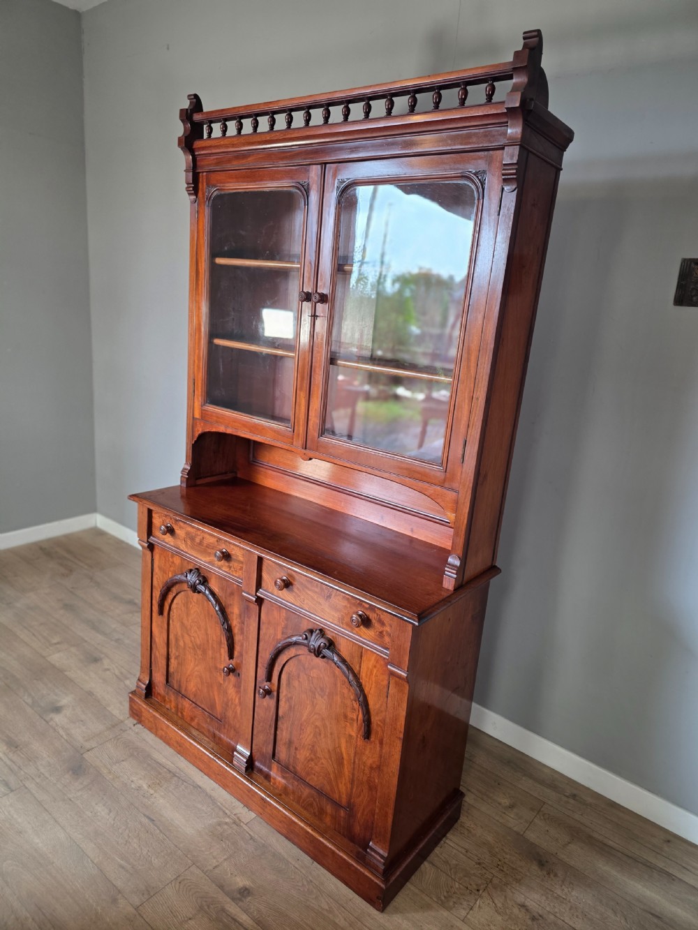 A Victorian mahogany glazed bookcase featuring a top spindle gallery, two glass cabinet doors, two drawers, and a lower double door cupboard on a plinth base. View VI