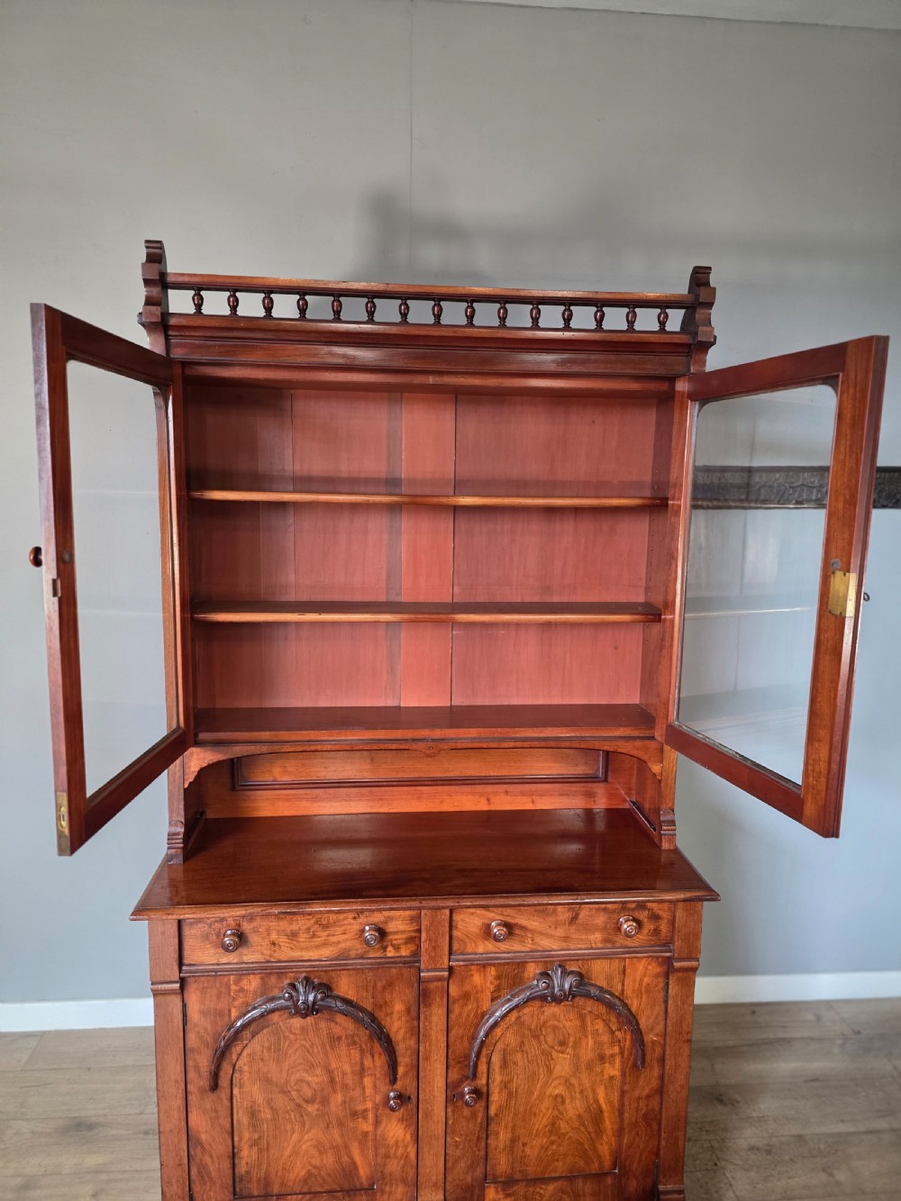 A Victorian mahogany glazed bookcase featuring a top spindle gallery, two glass cabinet doors, two drawers, and a lower double door cupboard on a plinth base. View XV