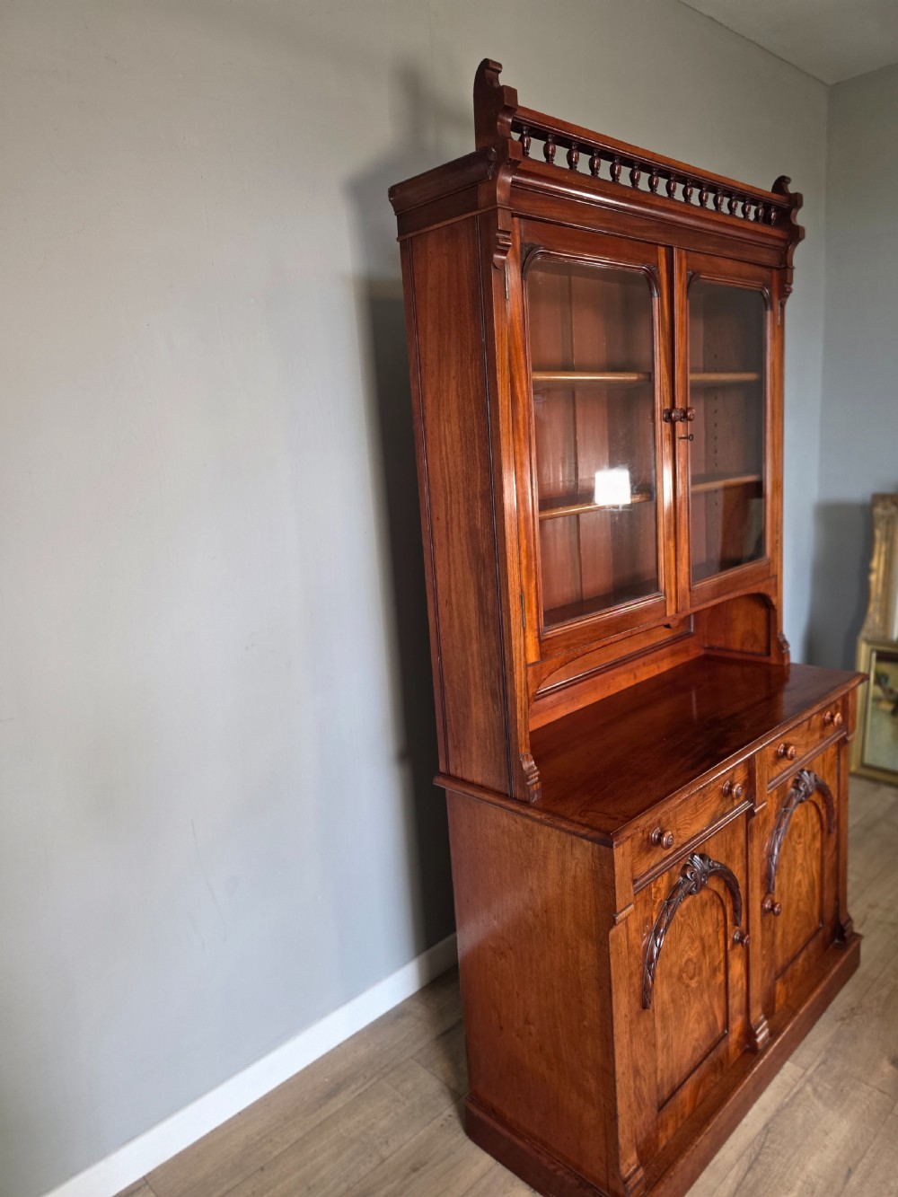 A Victorian mahogany glazed bookcase featuring a top spindle gallery, two glass cabinet doors, two drawers, and a lower double door cupboard on a plinth base. View XX