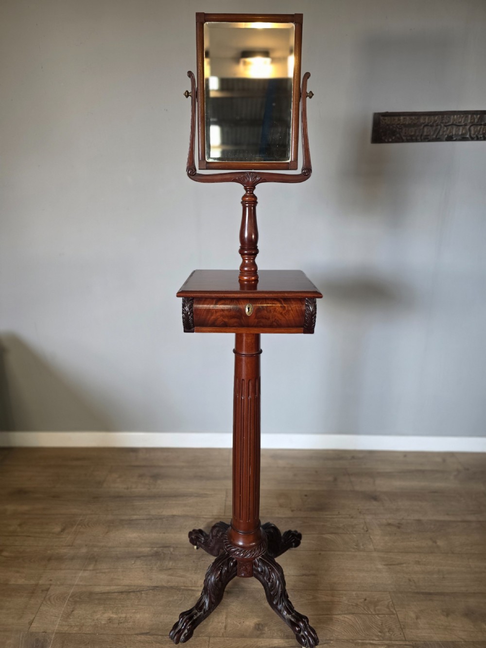 A tall mahogany gentlemans shaving stand featuring an adjustable rectangular mirror, a single drawer pedestal, a fluted column, and a carved tripod base.