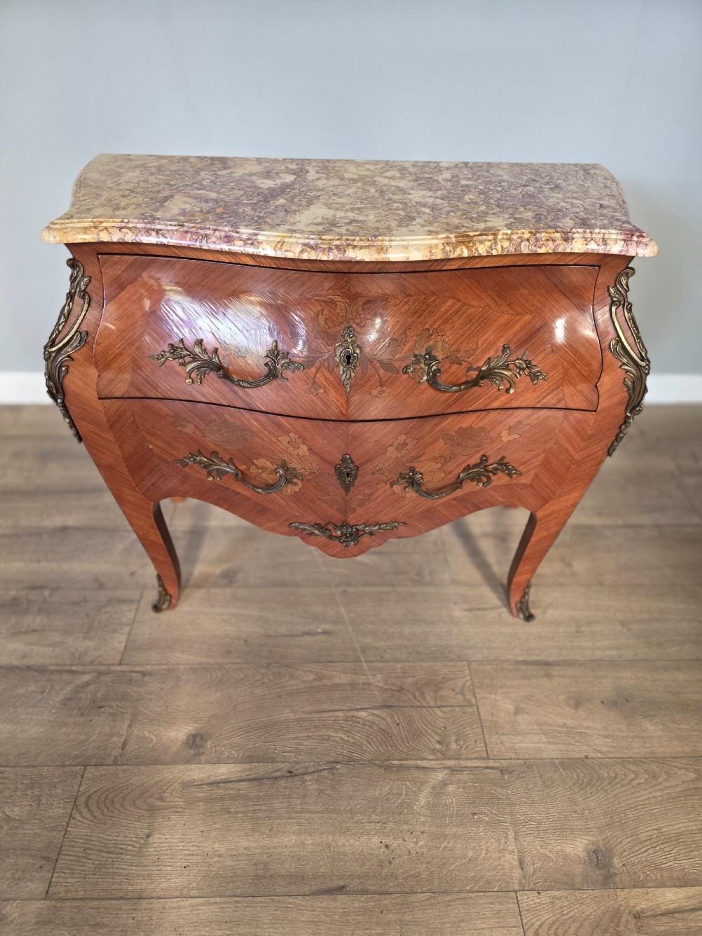 A French Rococo style bombe commode featuring a shaped marble top, floral marquetry veneer, two drawers, and ornamental gilt bronze mounts.