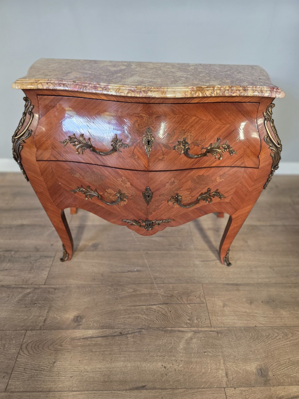 A French Rococo style bombe commode featuring a shaped marble top, floral marquetry veneer, two drawers, and ornamental gilt bronze mounts. View II