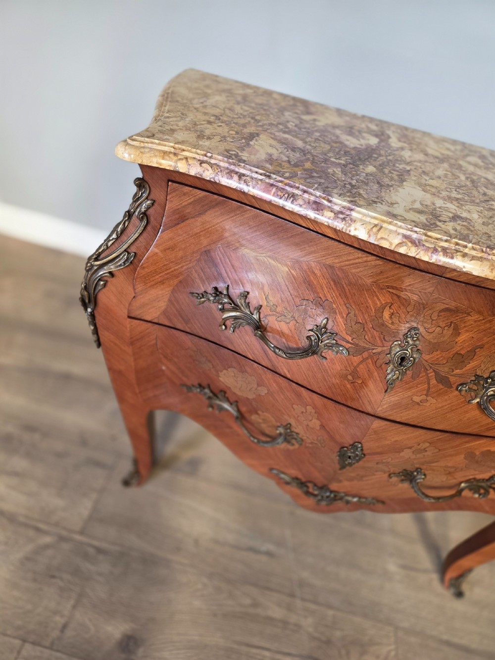 A French Rococo style bombe commode featuring a shaped marble top, floral marquetry veneer, two drawers, and ornamental gilt bronze mounts. View V
