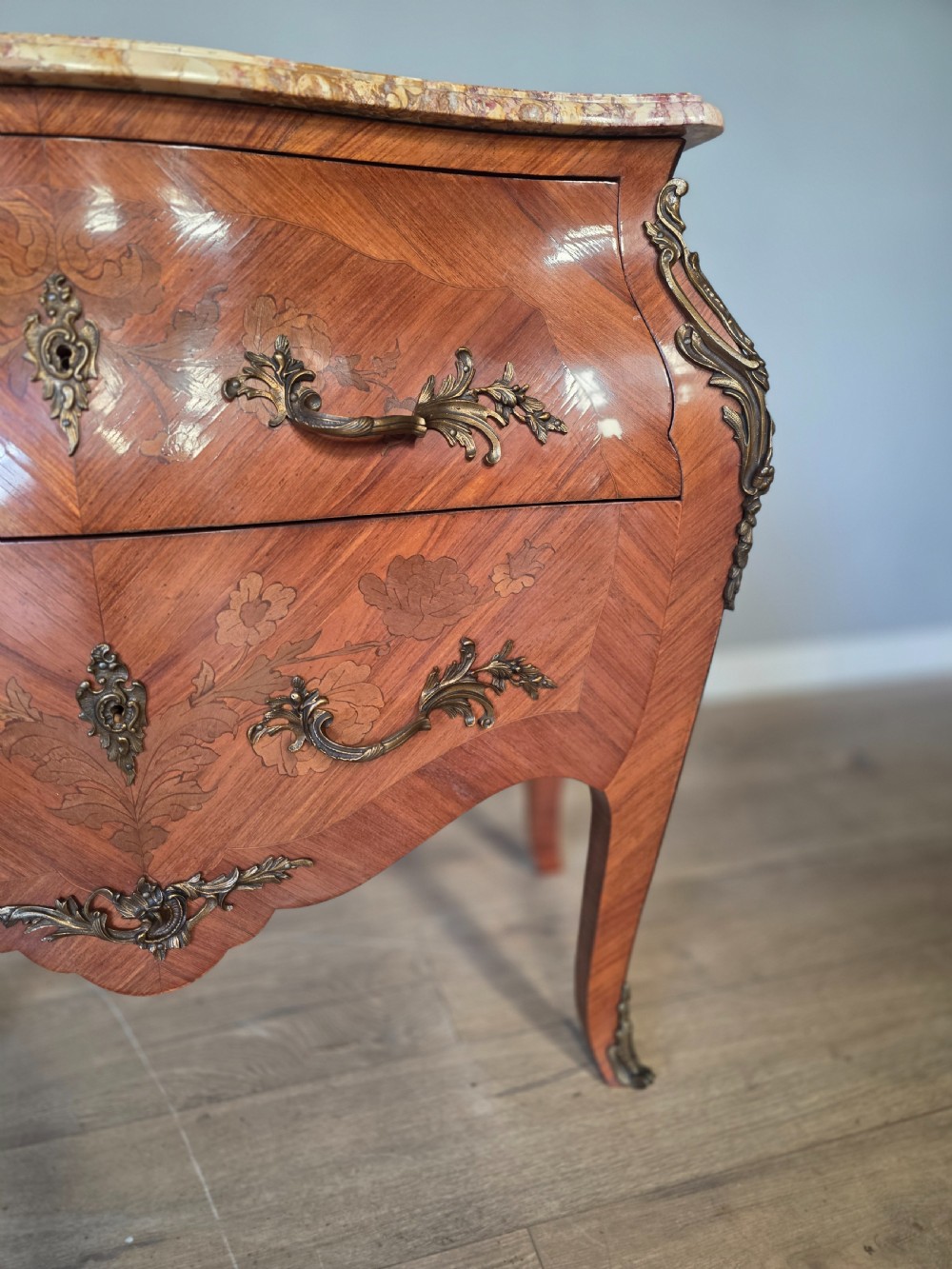 A French Rococo style bombe commode featuring a shaped marble top, floral marquetry veneer, two drawers, and ornamental gilt bronze mounts. View X