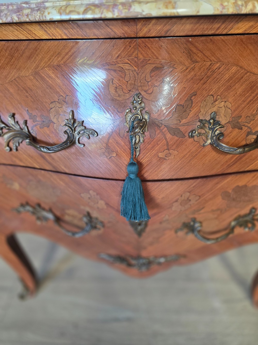 A French Rococo style bombe commode featuring a shaped marble top, floral marquetry veneer, two drawers, and ornamental gilt bronze mounts. View XIV