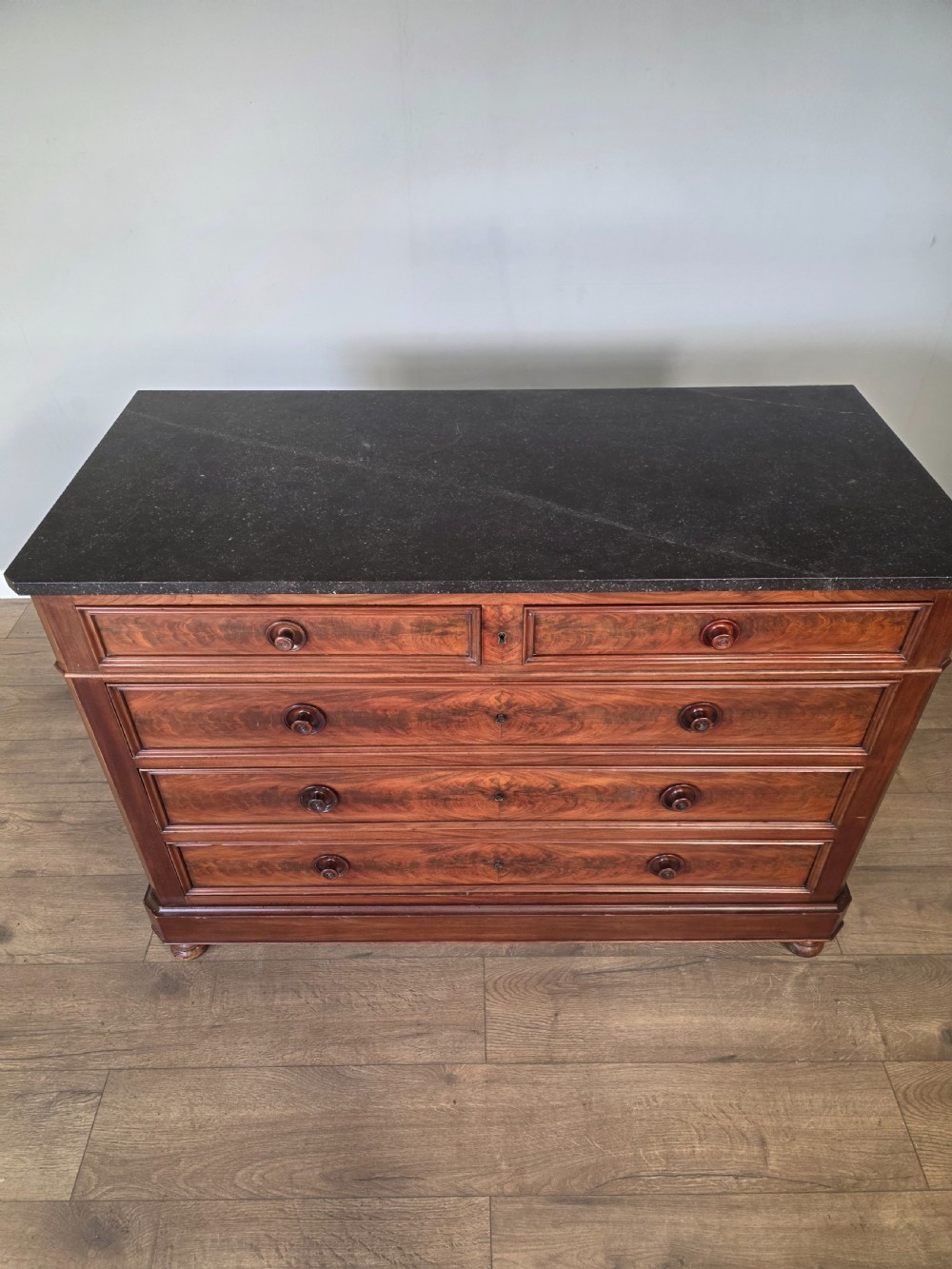 A nineteenth-century French mahogany chest of drawers featuring a dark marble top, flame mahogany drawer fronts, turned wooden knobs, and a plinth base. View V