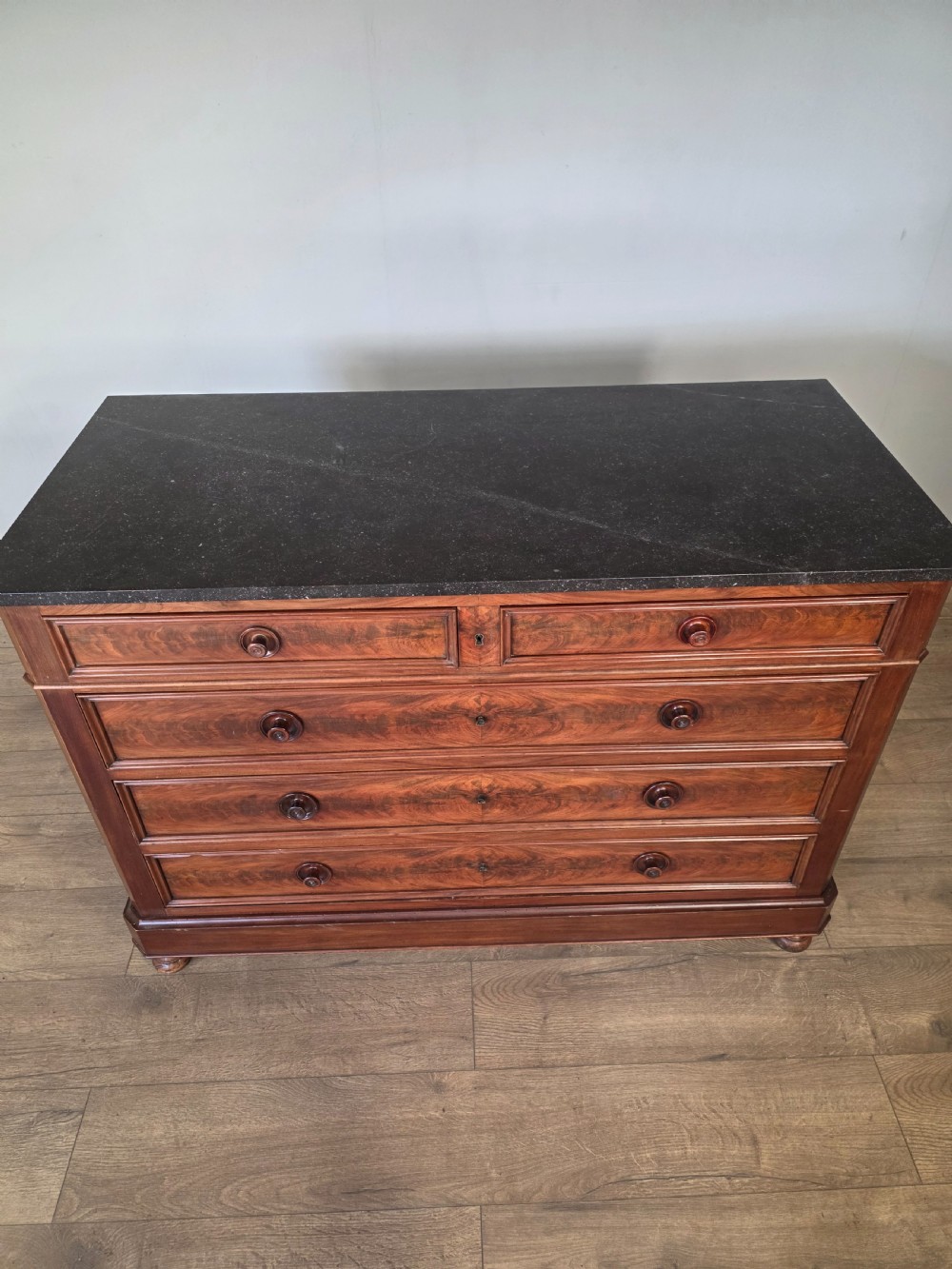 A nineteenth-century French mahogany chest of drawers featuring a dark marble top, flame mahogany drawer fronts, turned wooden knobs, and a plinth base. View VI