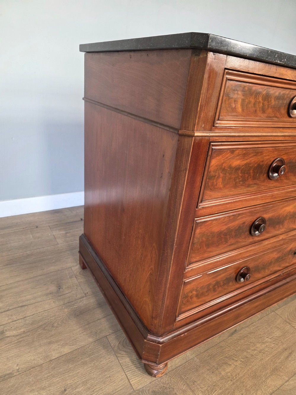 A nineteenth-century French mahogany chest of drawers featuring a dark marble top, flame mahogany drawer fronts, turned wooden knobs, and a plinth base. View X
