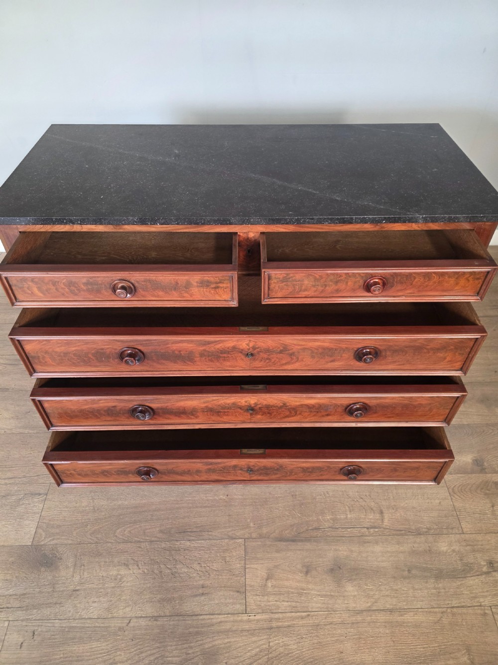 A nineteenth-century French mahogany chest of drawers featuring a dark marble top, flame mahogany drawer fronts, turned wooden knobs, and a plinth base. View XI