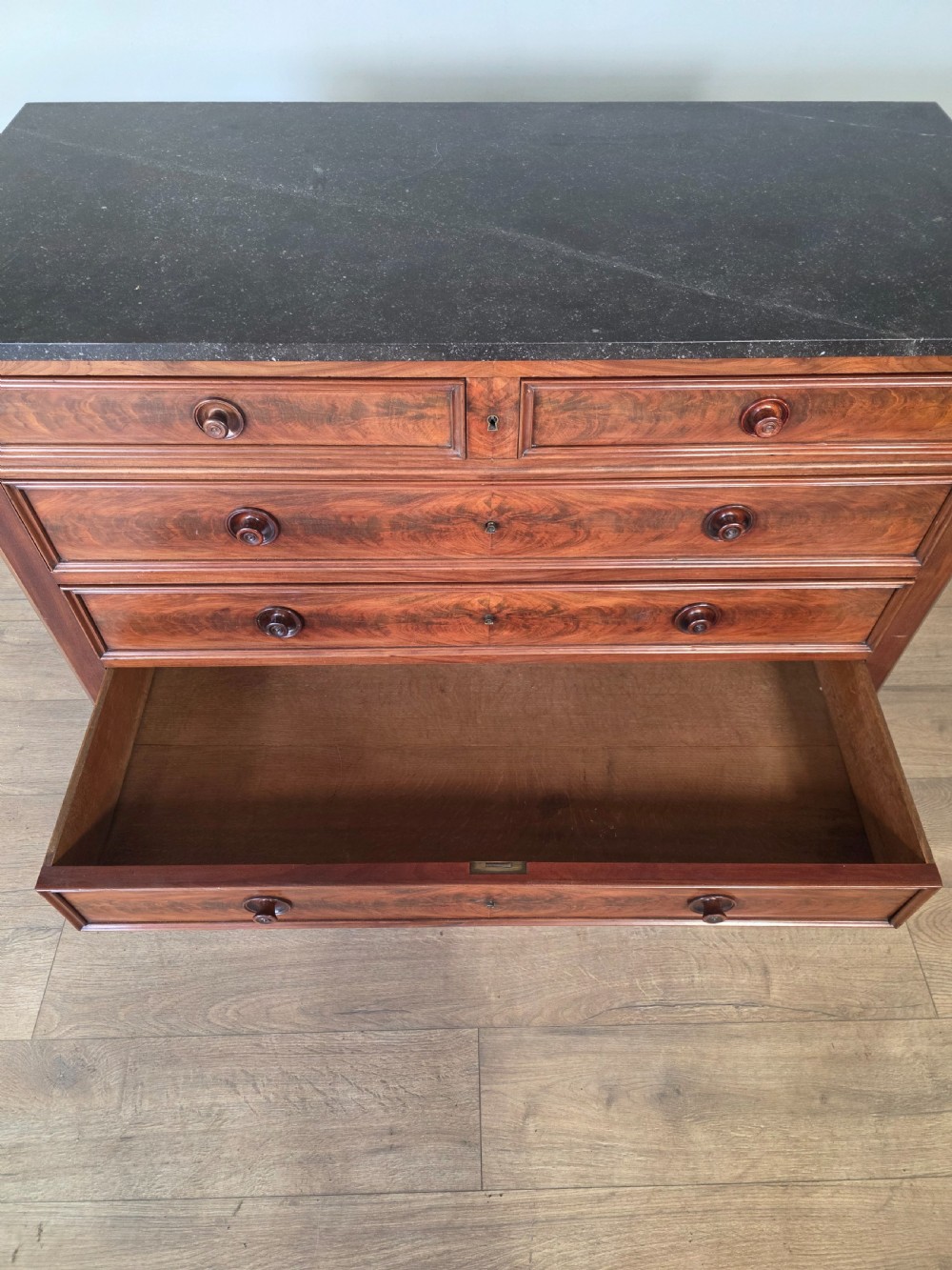 A nineteenth-century French mahogany chest of drawers featuring a dark marble top, flame mahogany drawer fronts, turned wooden knobs, and a plinth base. View XII