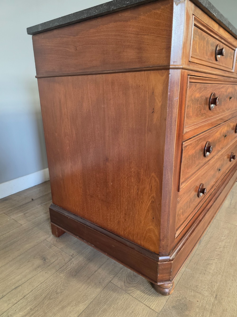 A nineteenth-century French mahogany chest of drawers featuring a dark marble top, flame mahogany drawer fronts, turned wooden knobs, and a plinth base. View XVI