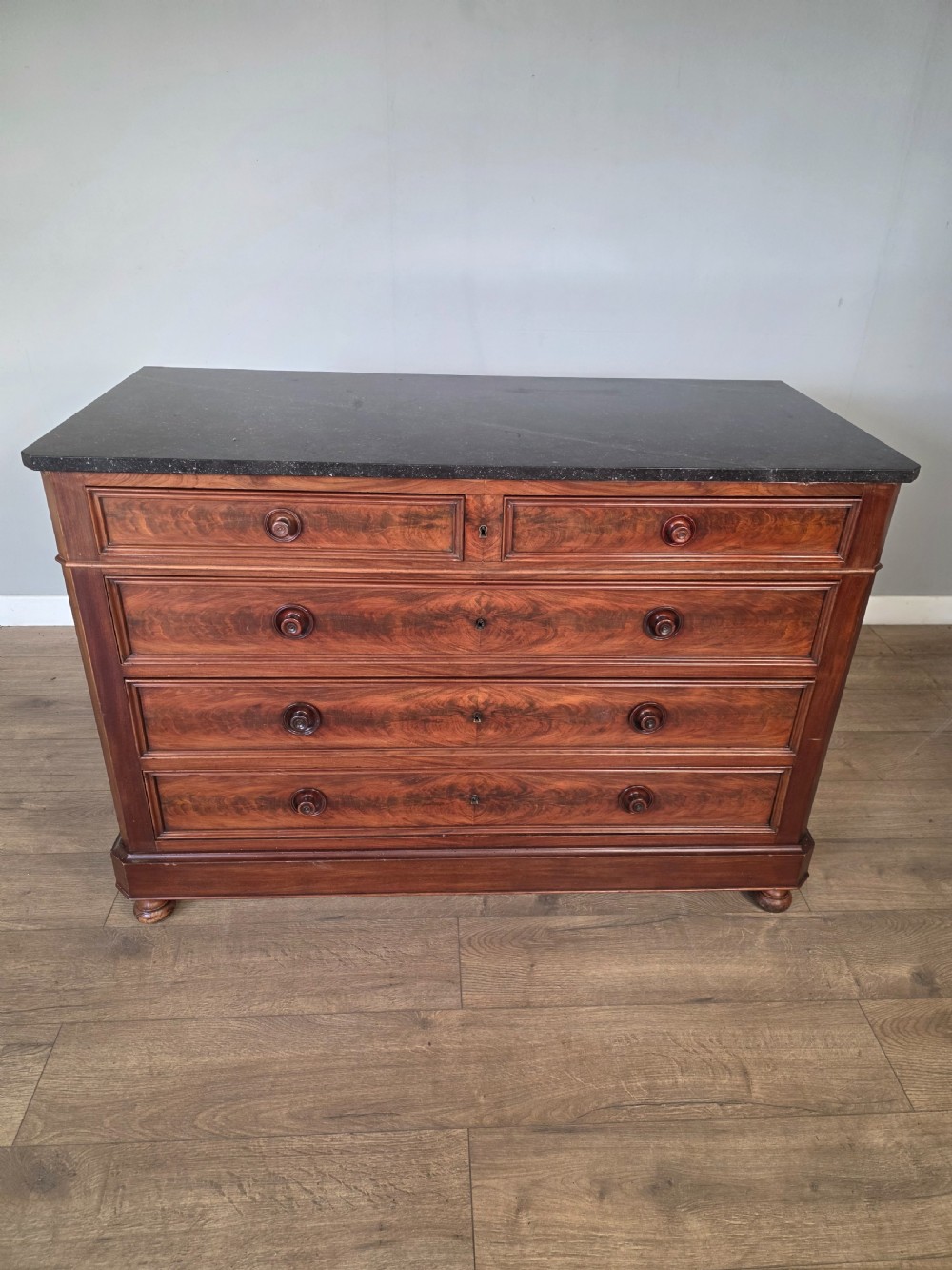 A nineteenth-century French mahogany chest of drawers featuring a dark marble top, flame mahogany drawer fronts, turned wooden knobs, and a plinth base. View XVII