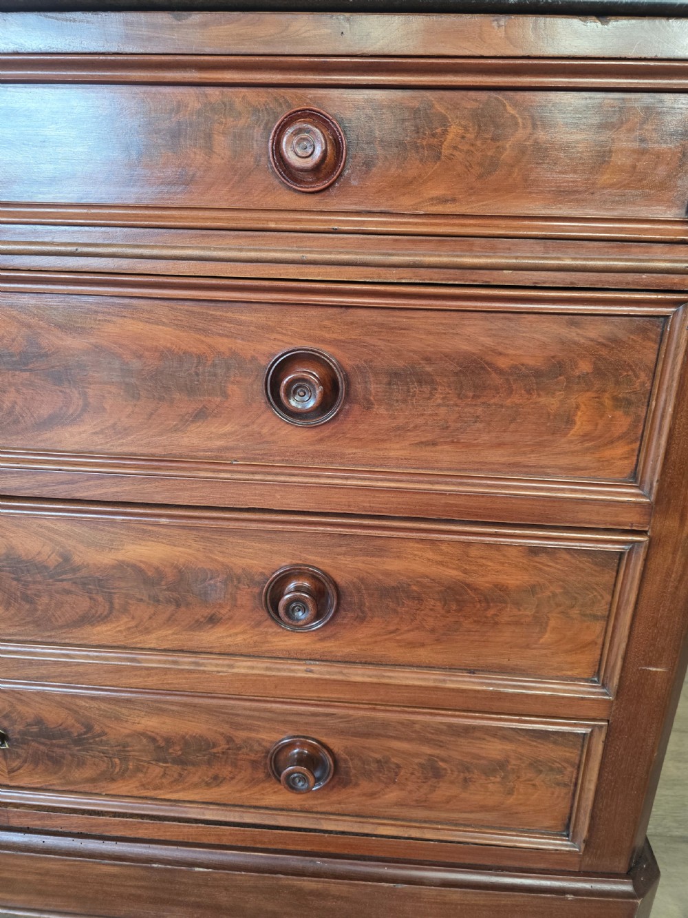 A nineteenth-century French mahogany chest of drawers featuring a dark marble top, flame mahogany drawer fronts, turned wooden knobs, and a plinth base. View XVIII