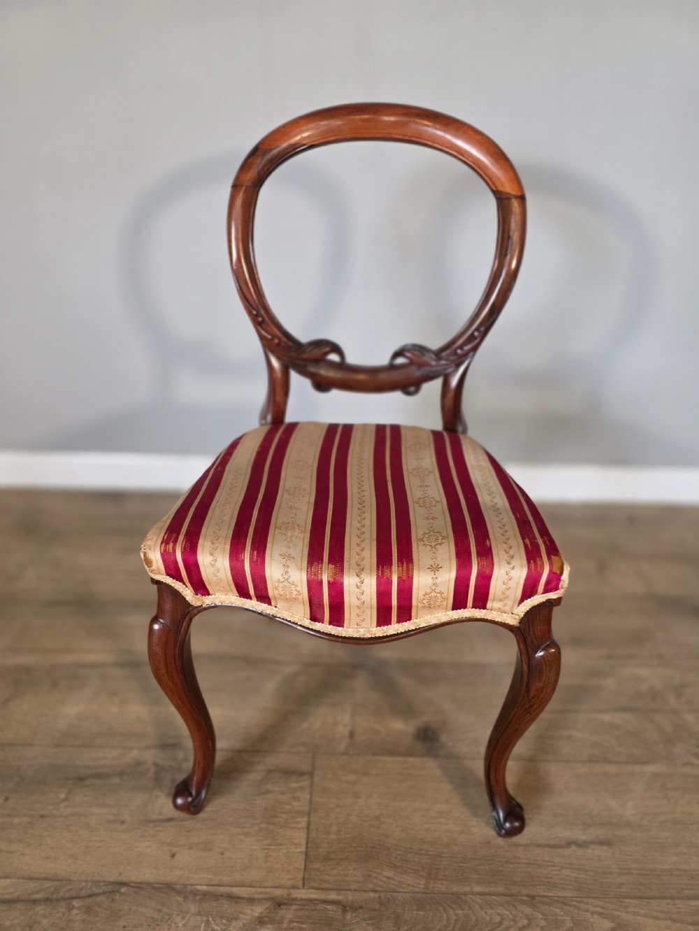 A set of six Victorian rosewood dining chairs featuring open balloon backs, carved waist rails, cabriole legs, and red and gold striped upholstered seats. View V