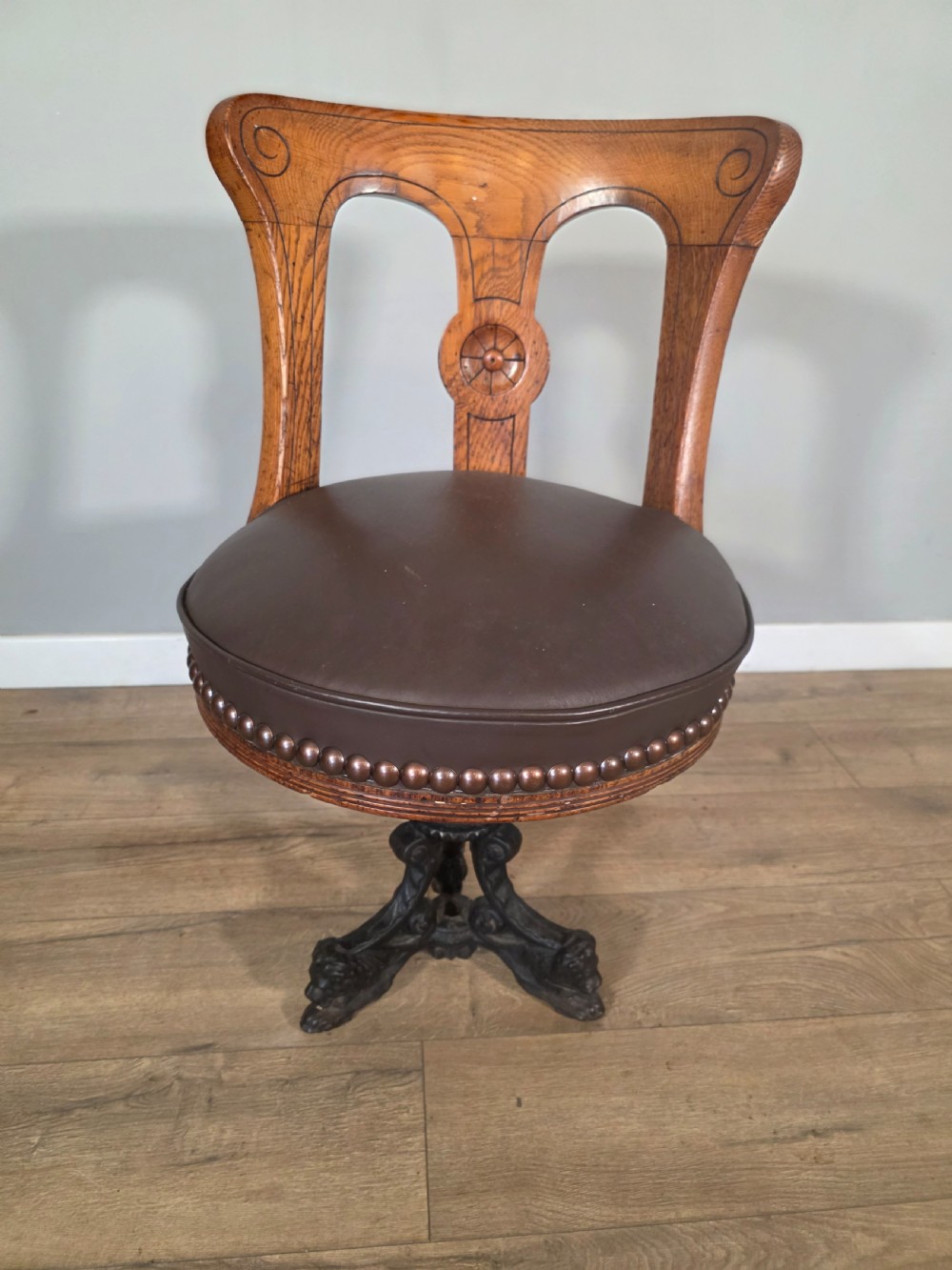 An antique 1890s swivel chair featuring a carved oak backrest, brown leather studded seat, and a cast iron pedestal base.