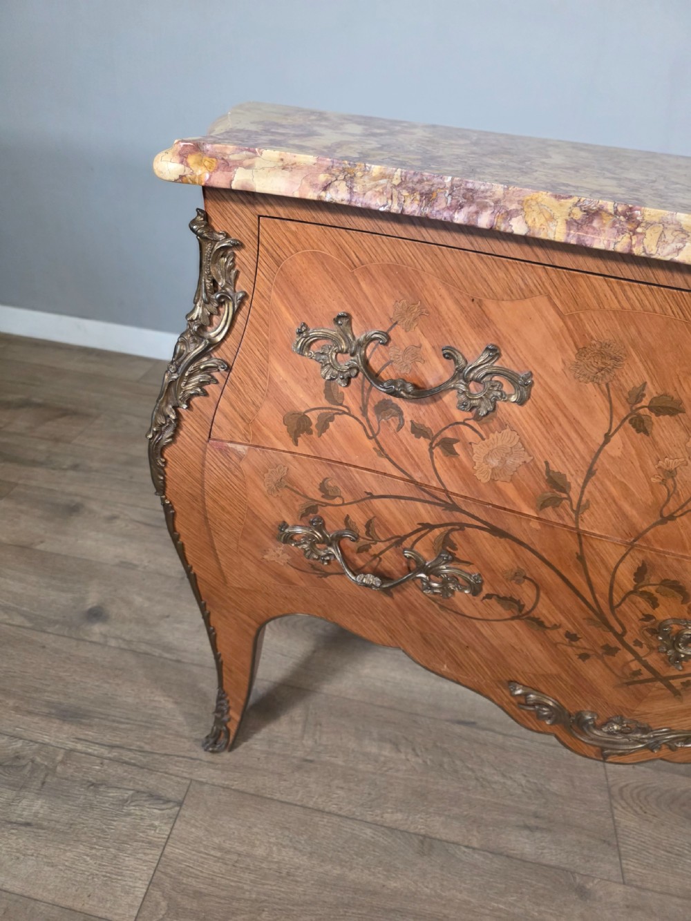 A French Louis XV style bombe commode featuring floral marquetry wood inlays, gilded metal mounts, cabriole legs, and a shaped pink-variegated marble top. View VI