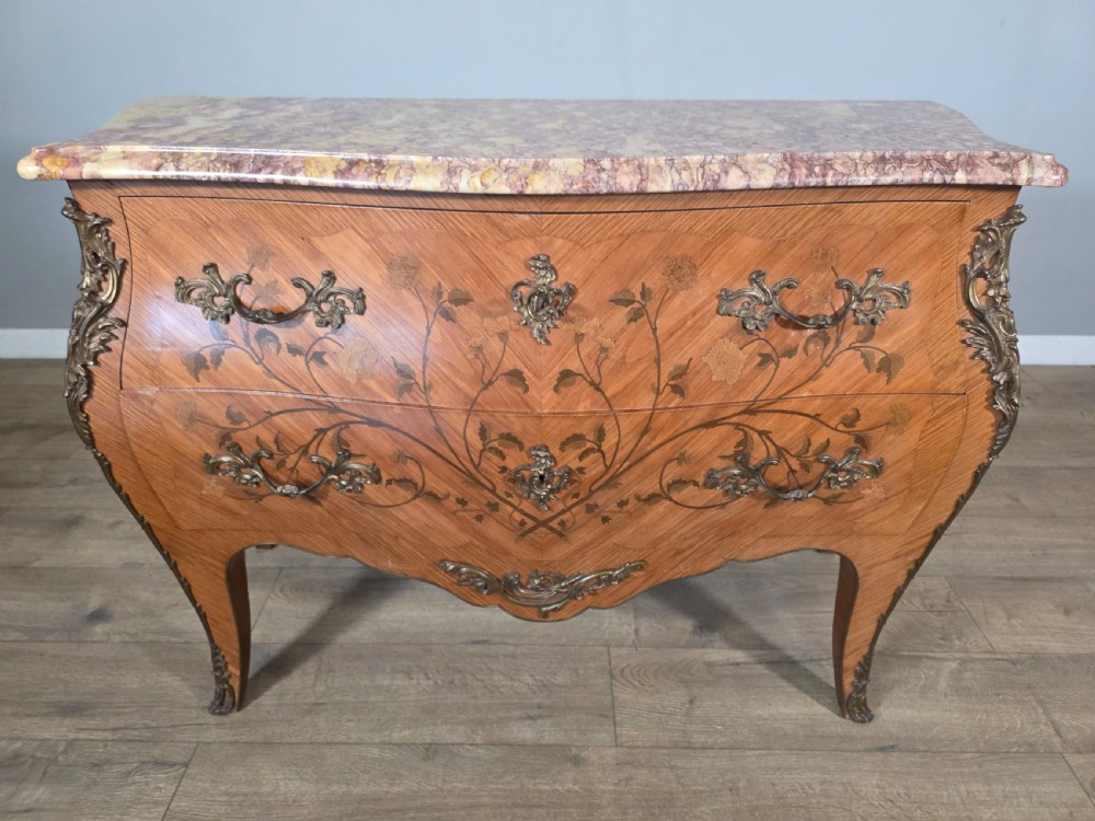 A French Louis XV style bombe commode featuring floral marquetry wood inlays, gilded metal mounts, cabriole legs, and a shaped pink-variegated marble top. View XV