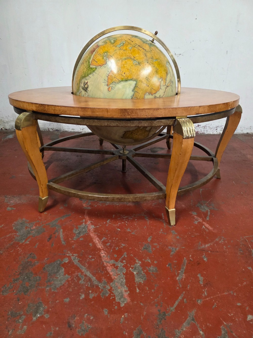 A circular mid-century coffee table featuring a central integrated terrestrial globe set within a honey brown wood veneer top, supported by curved legs and a brass framework. View V