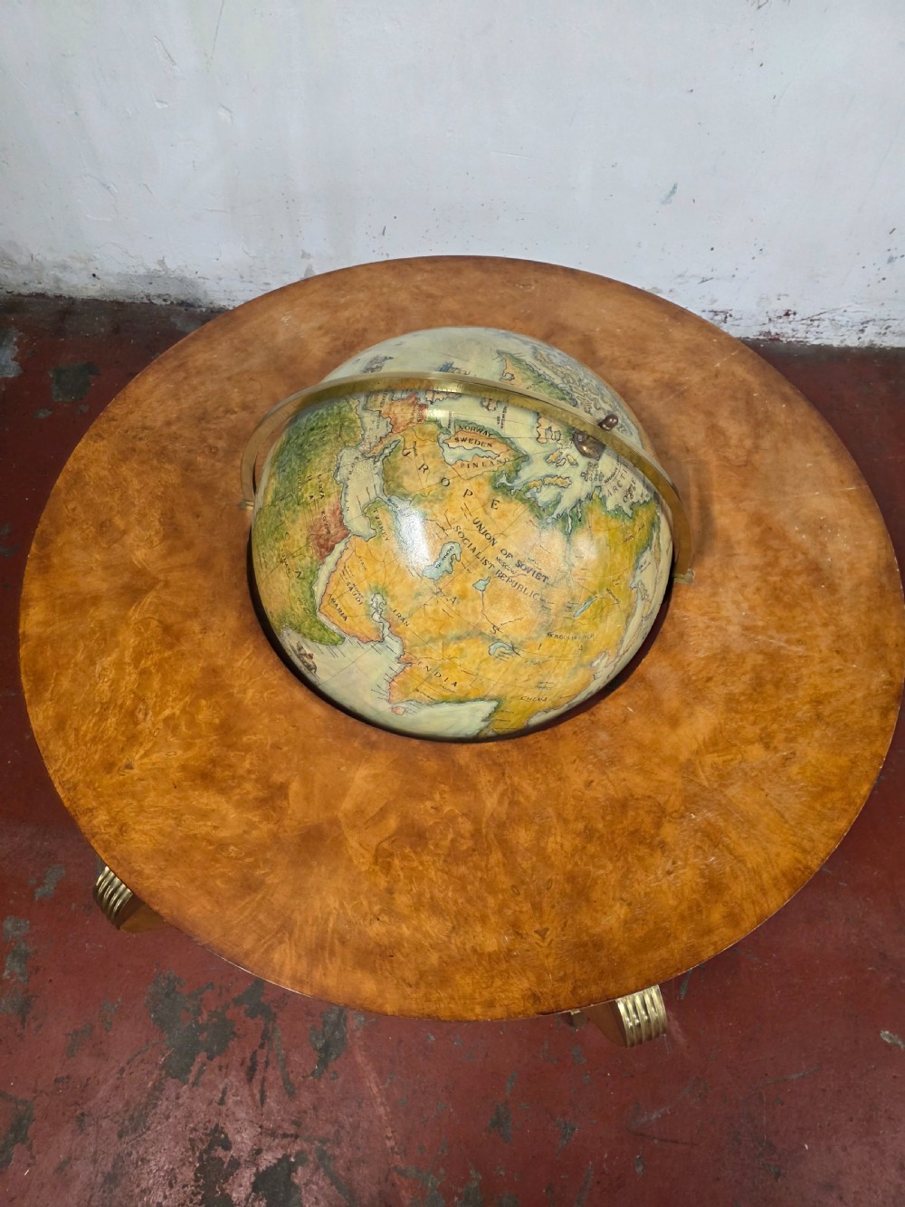 A circular mid-century coffee table featuring a central integrated terrestrial globe set within a honey brown wood veneer top, supported by curved legs and a brass framework. View VI