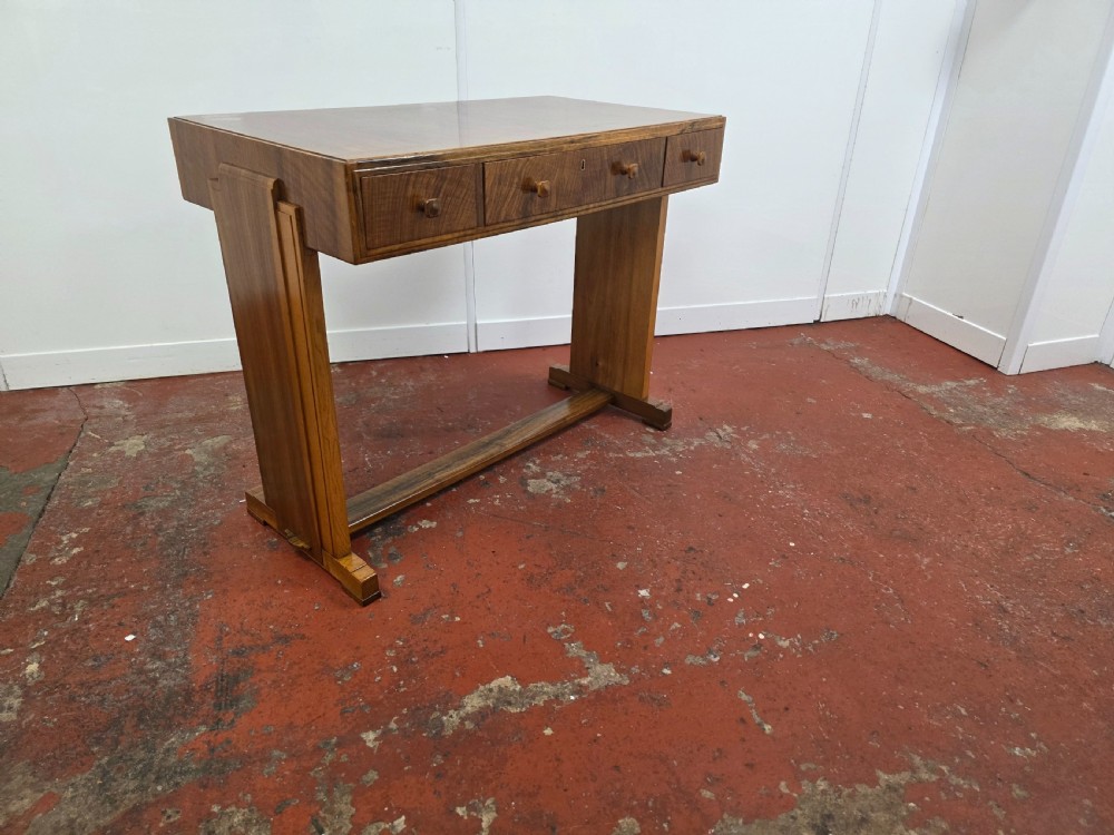 A 1930s Art Deco walnut desk featuring a rectangular top with three drawers, flat slab-style end legs, and a low cross stretcher.