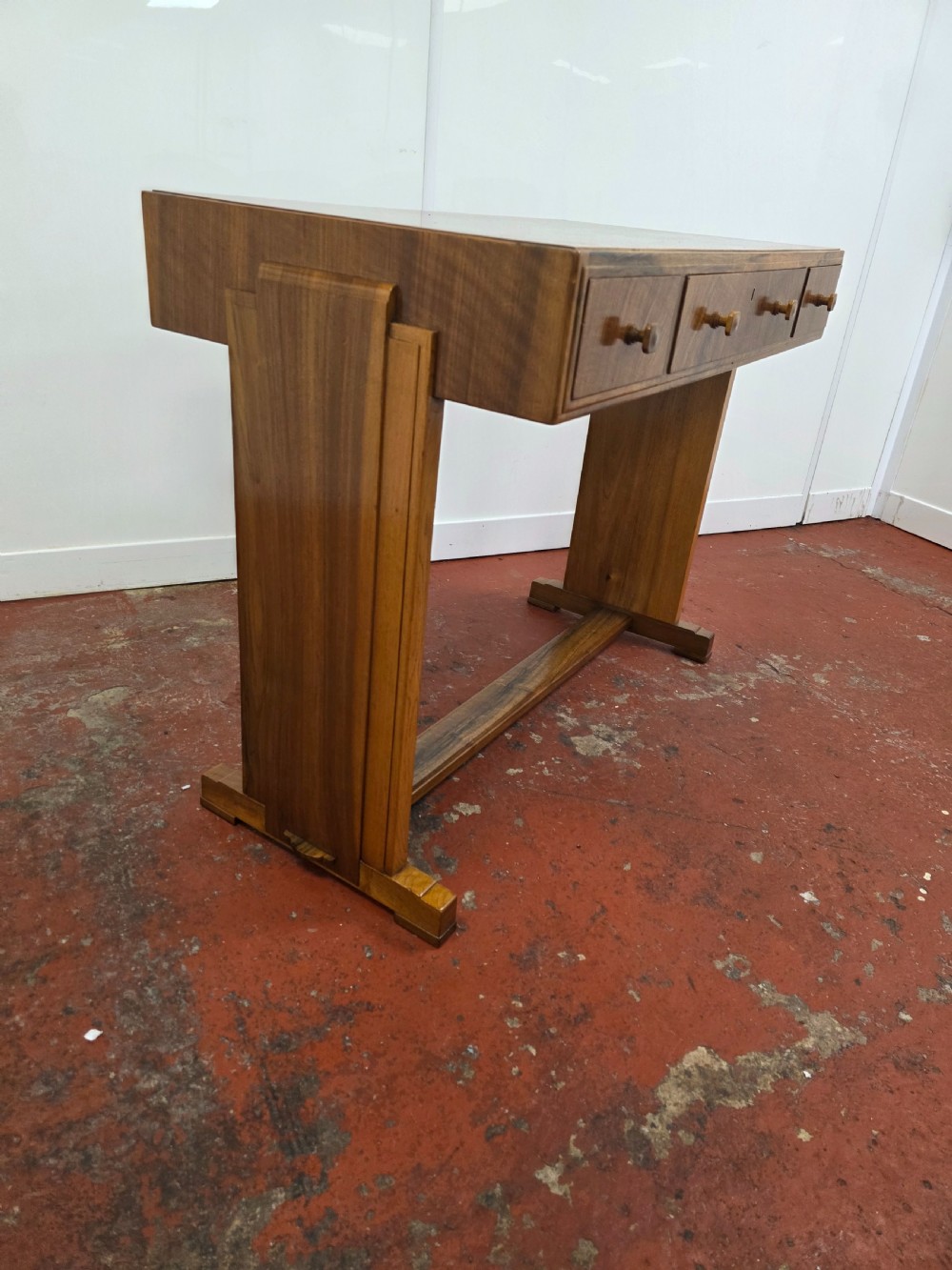 A 1930s Art Deco walnut desk featuring a rectangular top with three drawers, flat slab-style end legs, and a low cross stretcher. View V