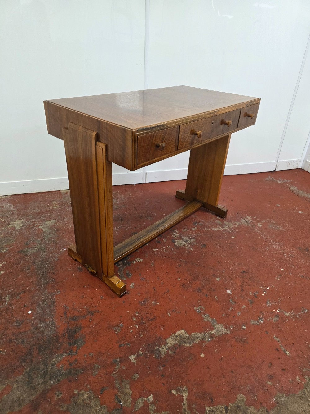 A 1930s Art Deco walnut desk featuring a rectangular top with three drawers, flat slab-style end legs, and a low cross stretcher. View VI