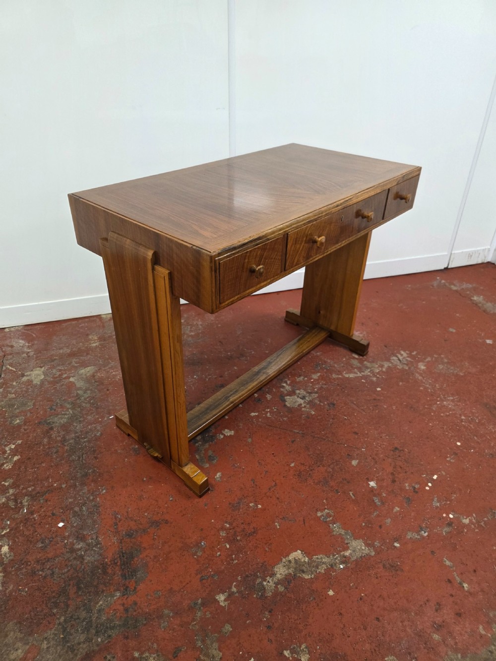 A 1930s Art Deco walnut desk featuring a rectangular top with three drawers, flat slab-style end legs, and a low cross stretcher. View VII