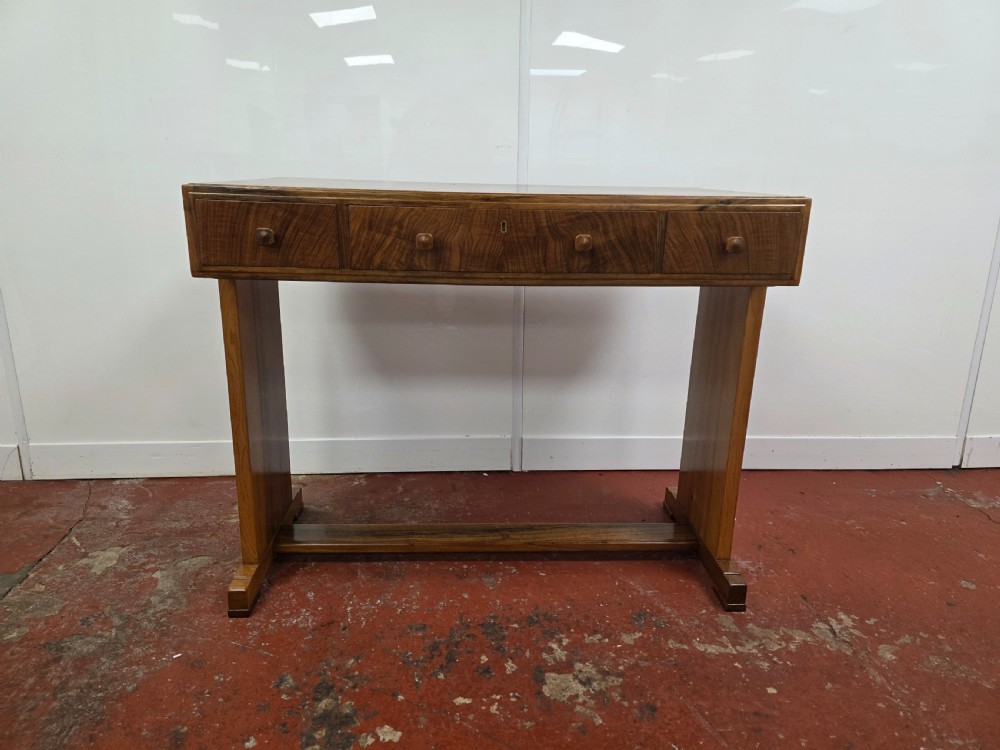 A 1930s Art Deco walnut desk featuring a rectangular top with three drawers, flat slab-style end legs, and a low cross stretcher. View X