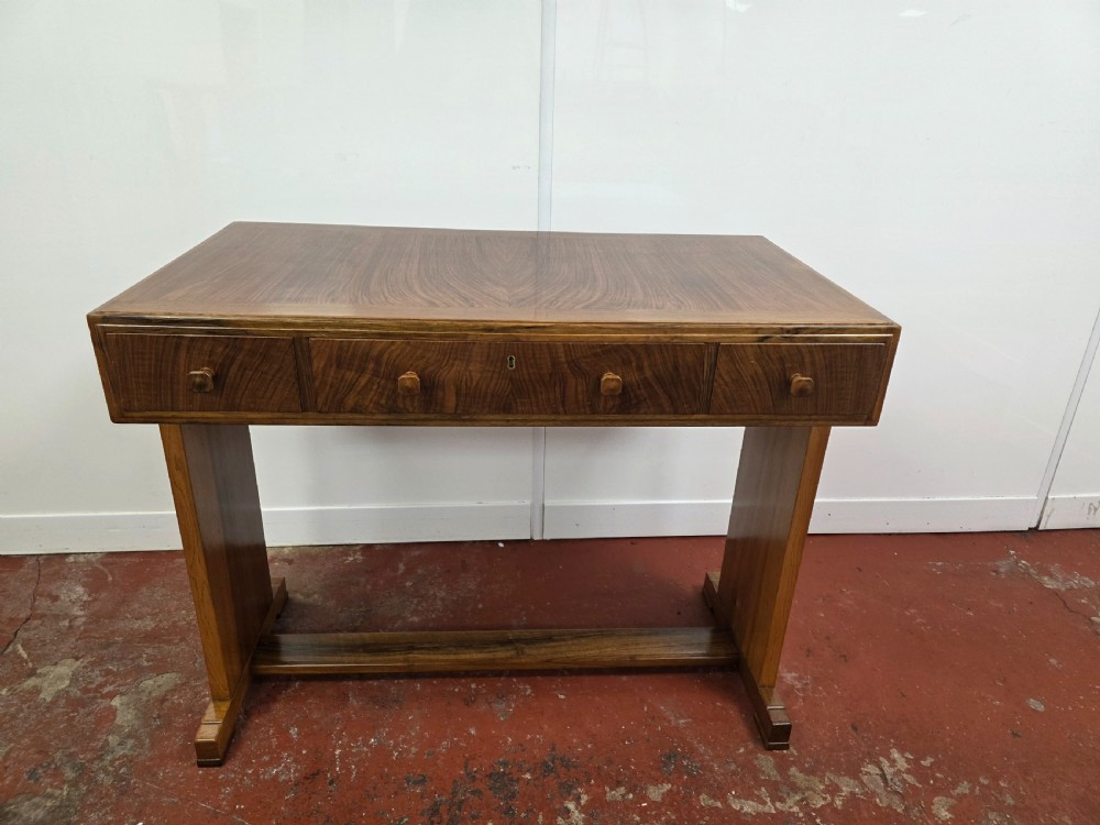A 1930s Art Deco walnut desk featuring a rectangular top with three drawers, flat slab-style end legs, and a low cross stretcher. View XI