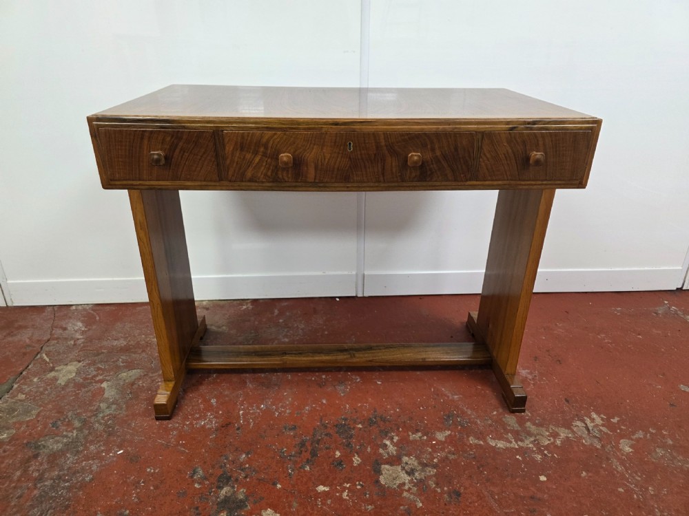 A 1930s Art Deco walnut desk featuring a rectangular top with three drawers, flat slab-style end legs, and a low cross stretcher. View XVII