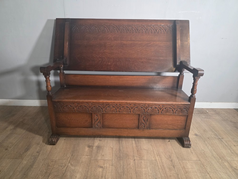 A 1920s carved dark oak monks bench featuring a high panelled backrest, turned arm supports, and a lower storage box base.