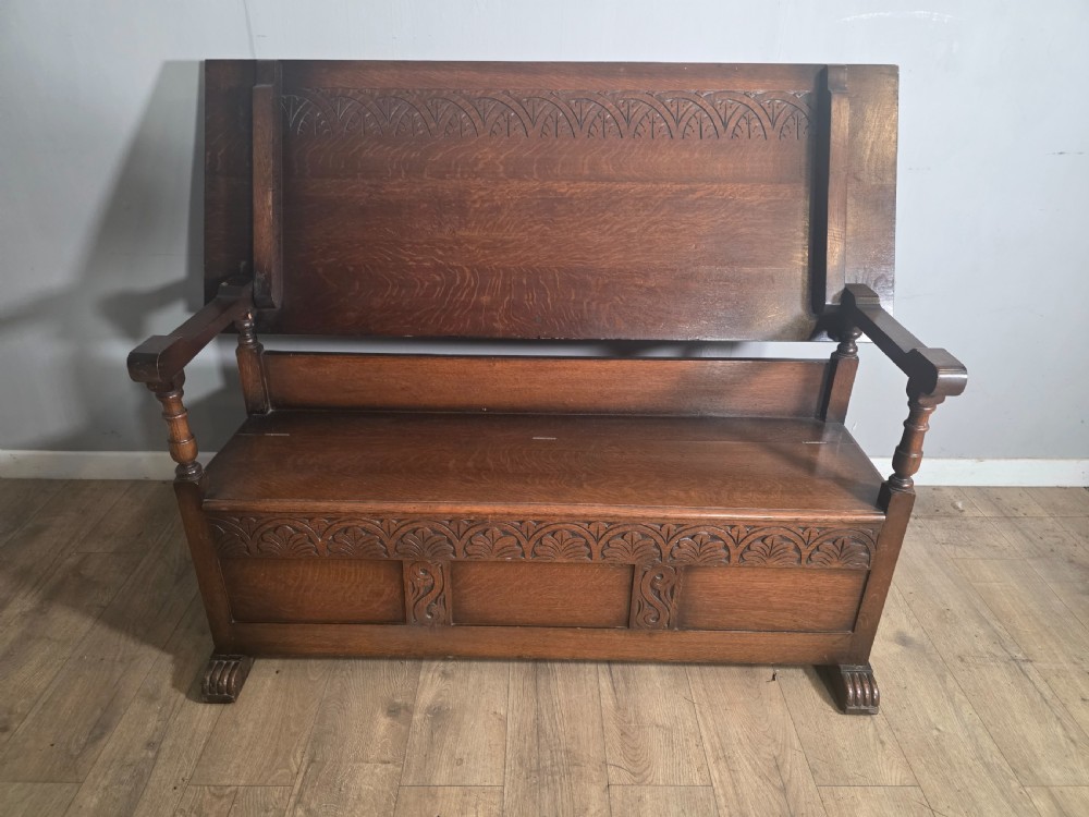 A 1920s carved dark oak monks bench featuring a high panelled backrest, turned arm supports, and a lower storage box base. View II