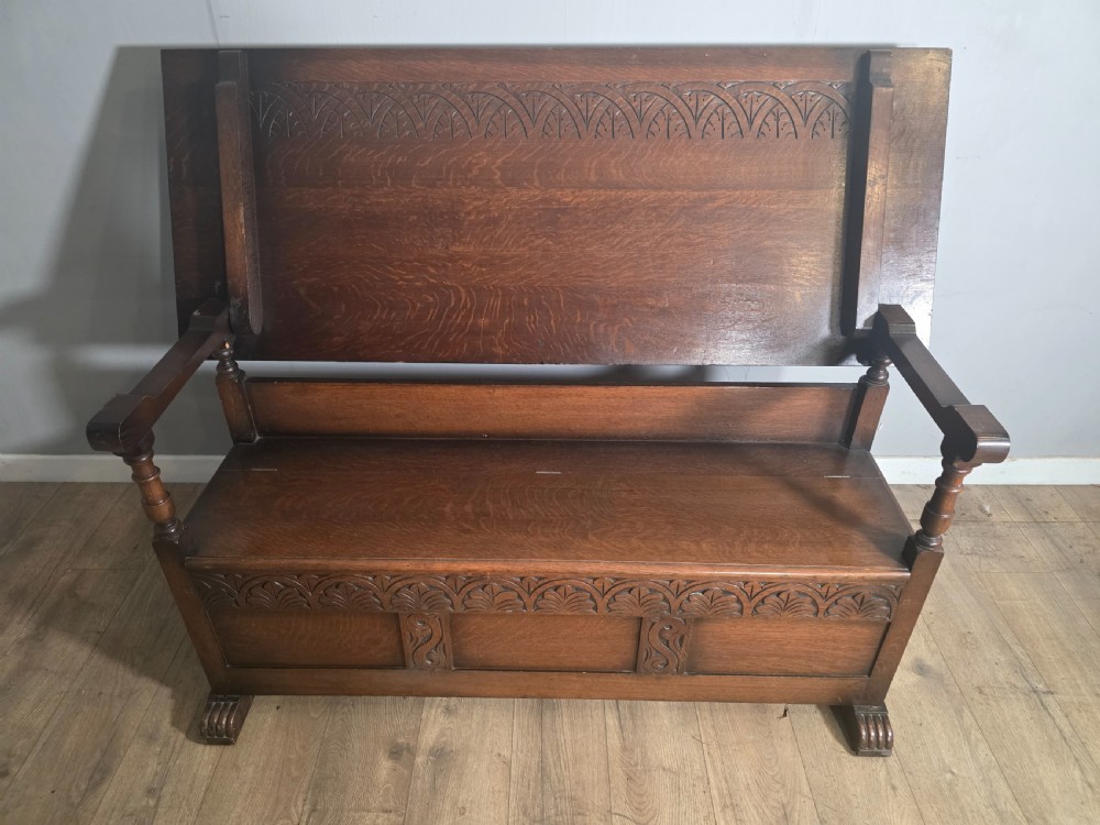 A 1920s carved dark oak monks bench featuring a high panelled backrest, turned arm supports, and a lower storage box base. View III