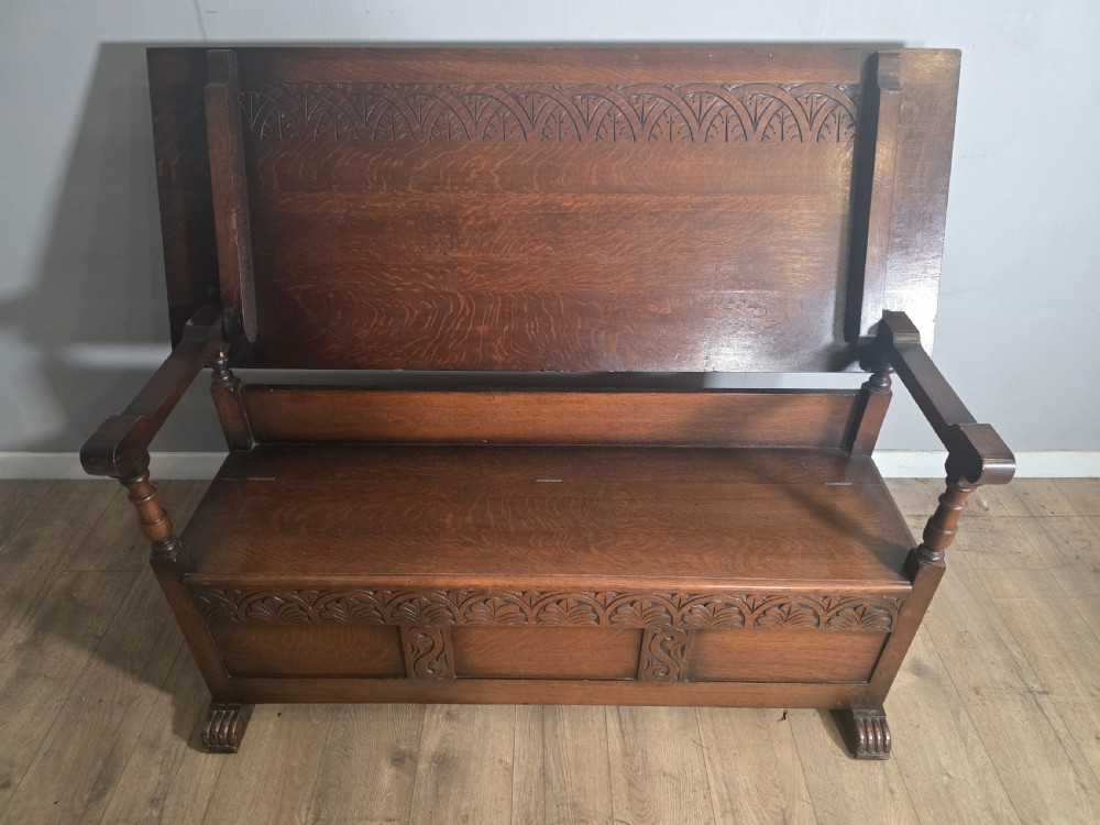A 1920s carved dark oak monks bench featuring a high panelled backrest, turned arm supports, and a lower storage box base. View IV