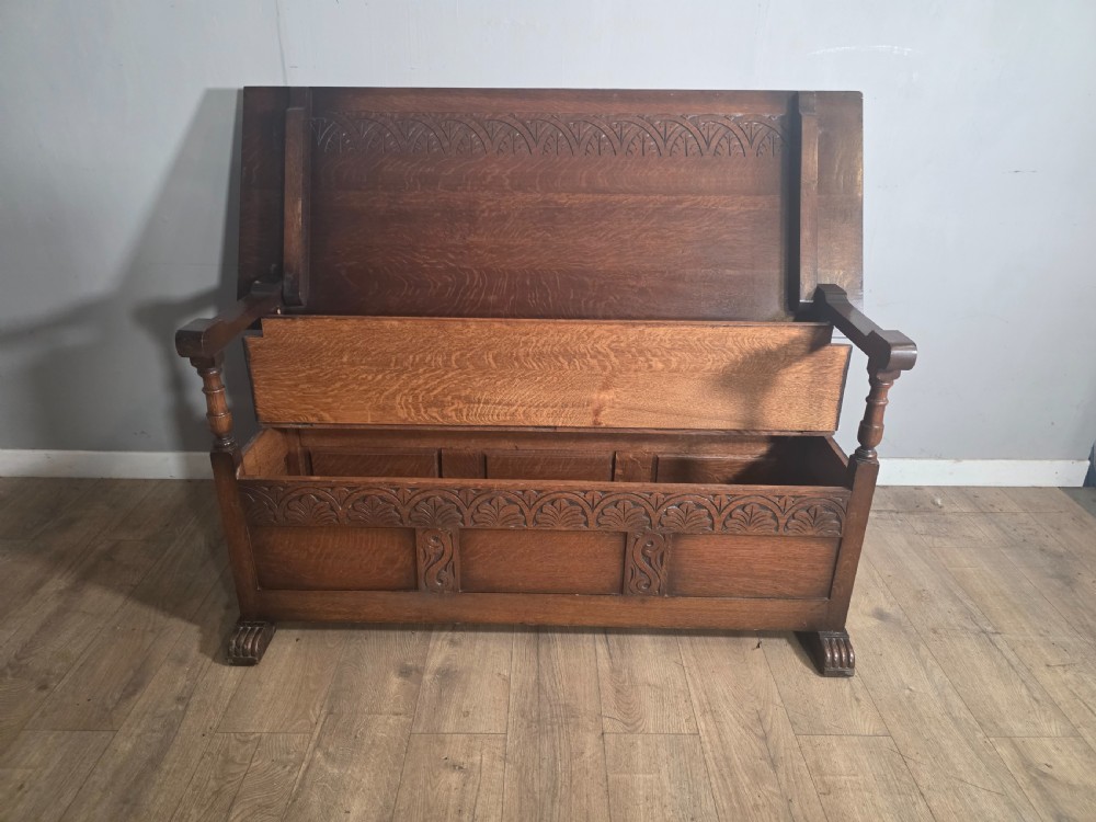 A 1920s carved dark oak monks bench featuring a high panelled backrest, turned arm supports, and a lower storage box base. View V