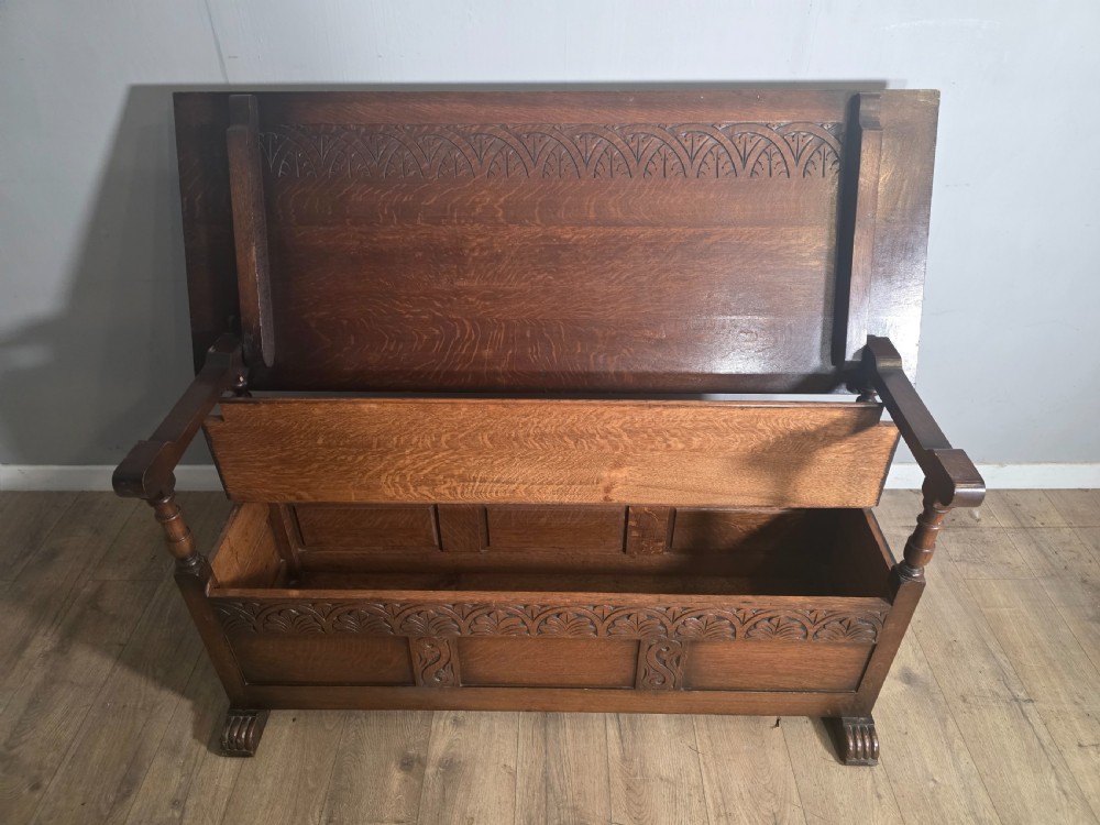 A 1920s carved dark oak monks bench featuring a high panelled backrest, turned arm supports, and a lower storage box base. View VI