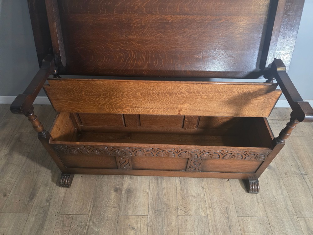 A 1920s carved dark oak monks bench featuring a high panelled backrest, turned arm supports, and a lower storage box base. View VII