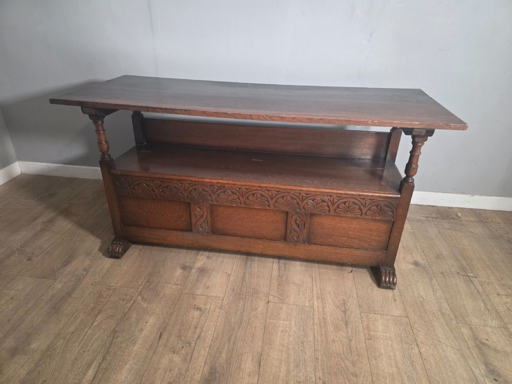 A 1920s carved dark oak monks bench featuring a high panelled backrest, turned arm supports, and a lower storage box base. View IX
