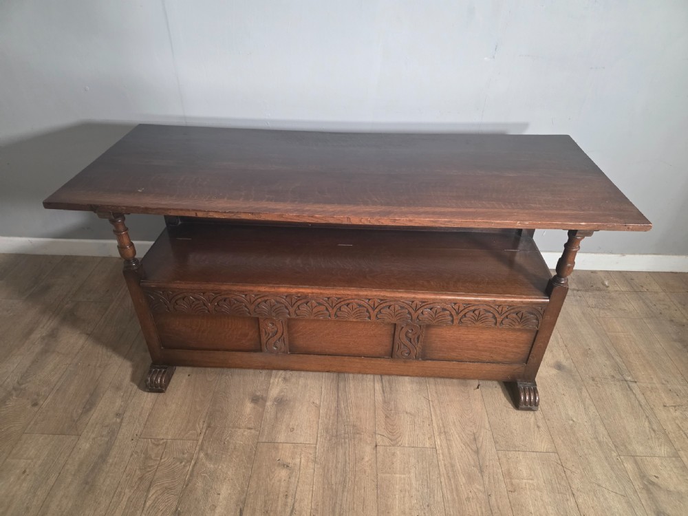 A 1920s carved dark oak monks bench featuring a high panelled backrest, turned arm supports, and a lower storage box base. View X