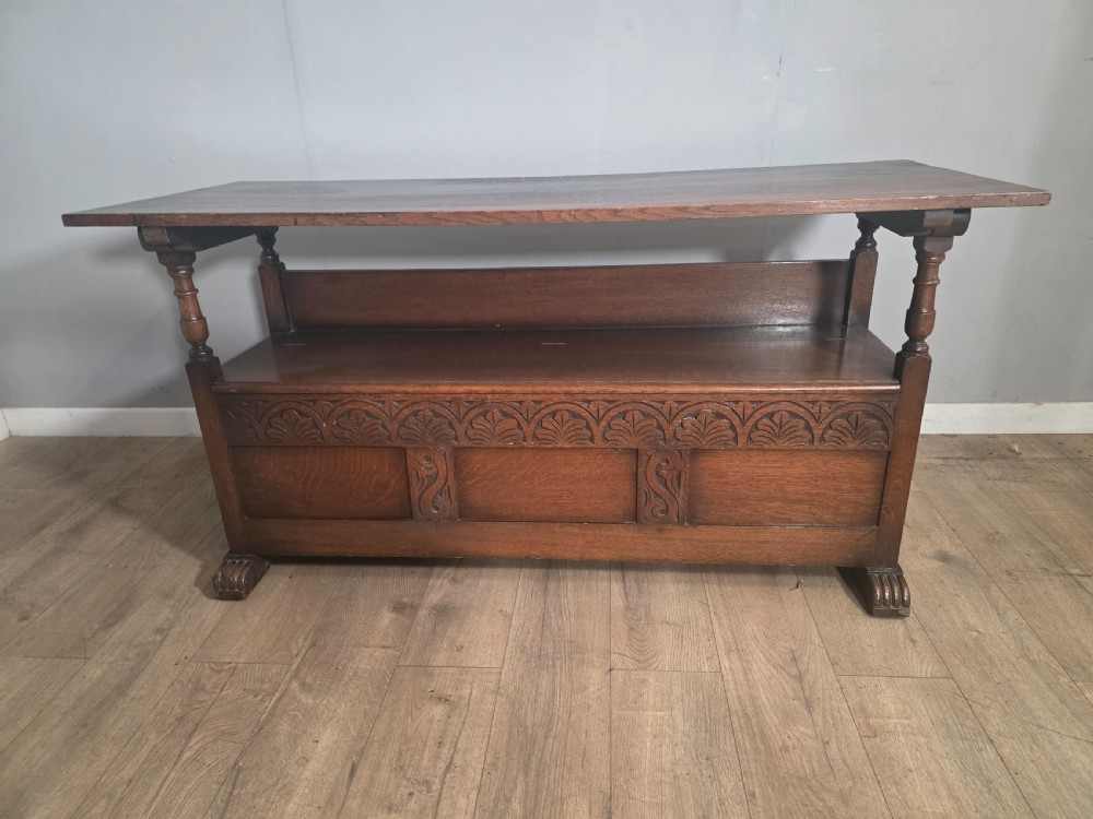 A 1920s carved dark oak monks bench featuring a high panelled backrest, turned arm supports, and a lower storage box base. View XVI