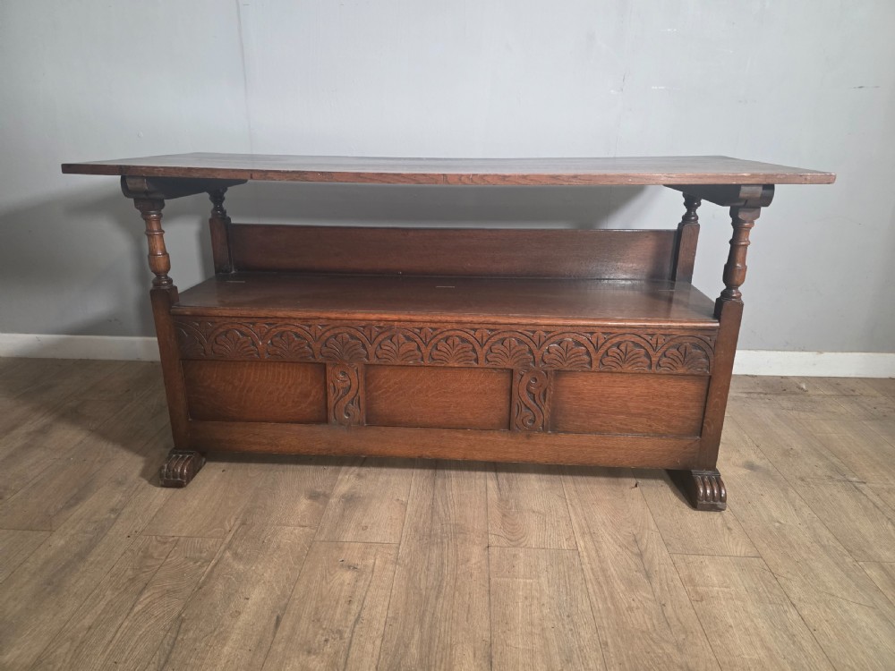 A 1920s carved dark oak monks bench featuring a high panelled backrest, turned arm supports, and a lower storage box base. View XVII