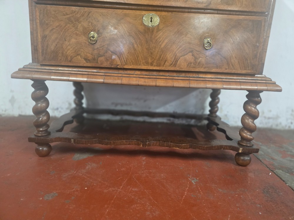 An early 18th-century Queen Anne burr walnut chest on stand featuring multiple graduated drawers with brass handles, resting on a base with barley twist legs and a shaped stretcher. View X