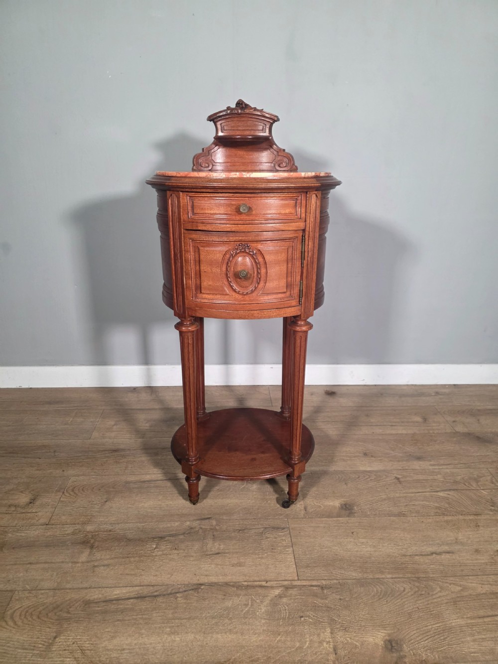 An antique French mahogany bedside cabinet featuring an oval marble top, a carved cresting gallery, a single drawer above a cupboard door, turned fluted legs, and a lower shelf.