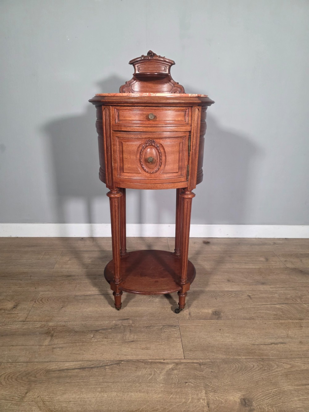 An antique French mahogany bedside cabinet featuring an oval marble top, a carved cresting gallery, a single drawer above a cupboard door, turned fluted legs, and a lower shelf. View II