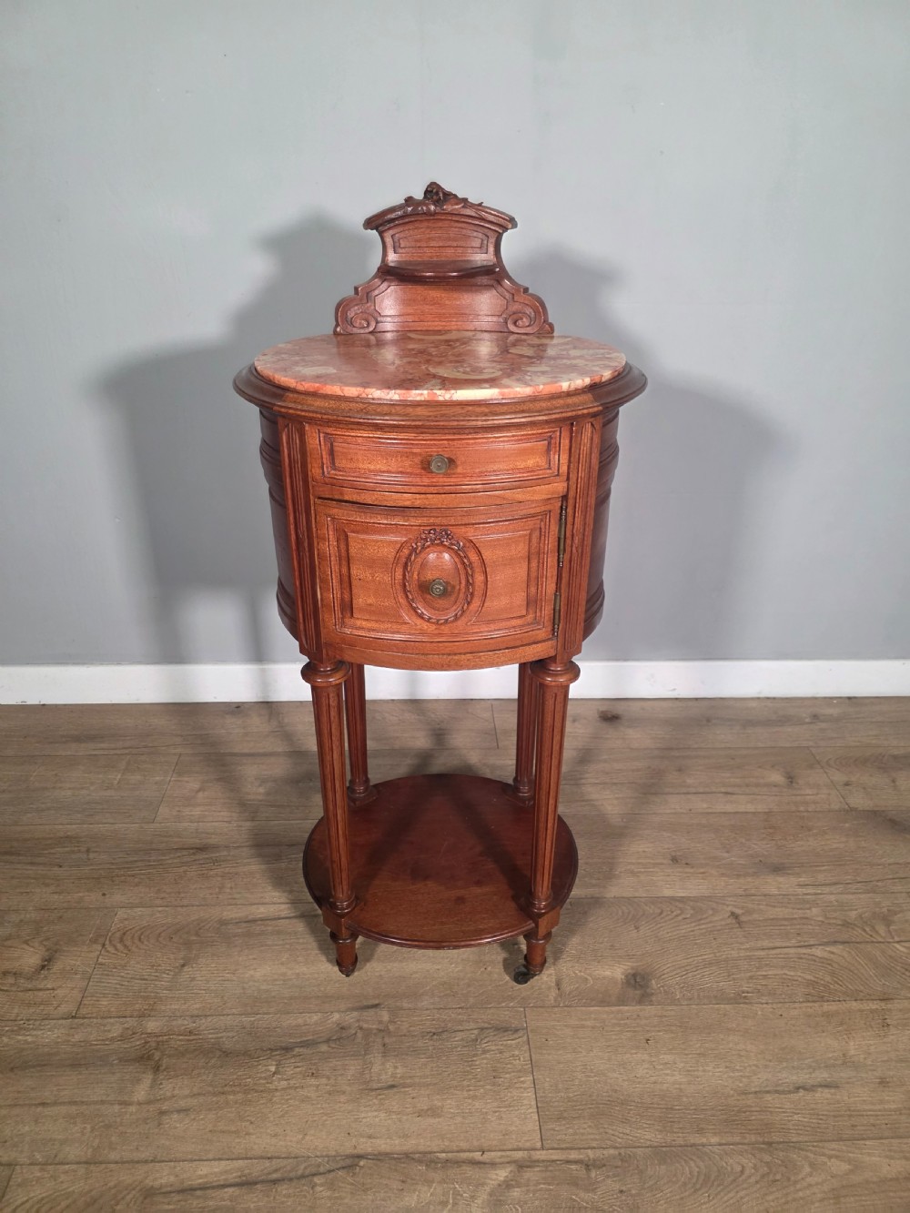 An antique French mahogany bedside cabinet featuring an oval marble top, a carved cresting gallery, a single drawer above a cupboard door, turned fluted legs, and a lower shelf. View III