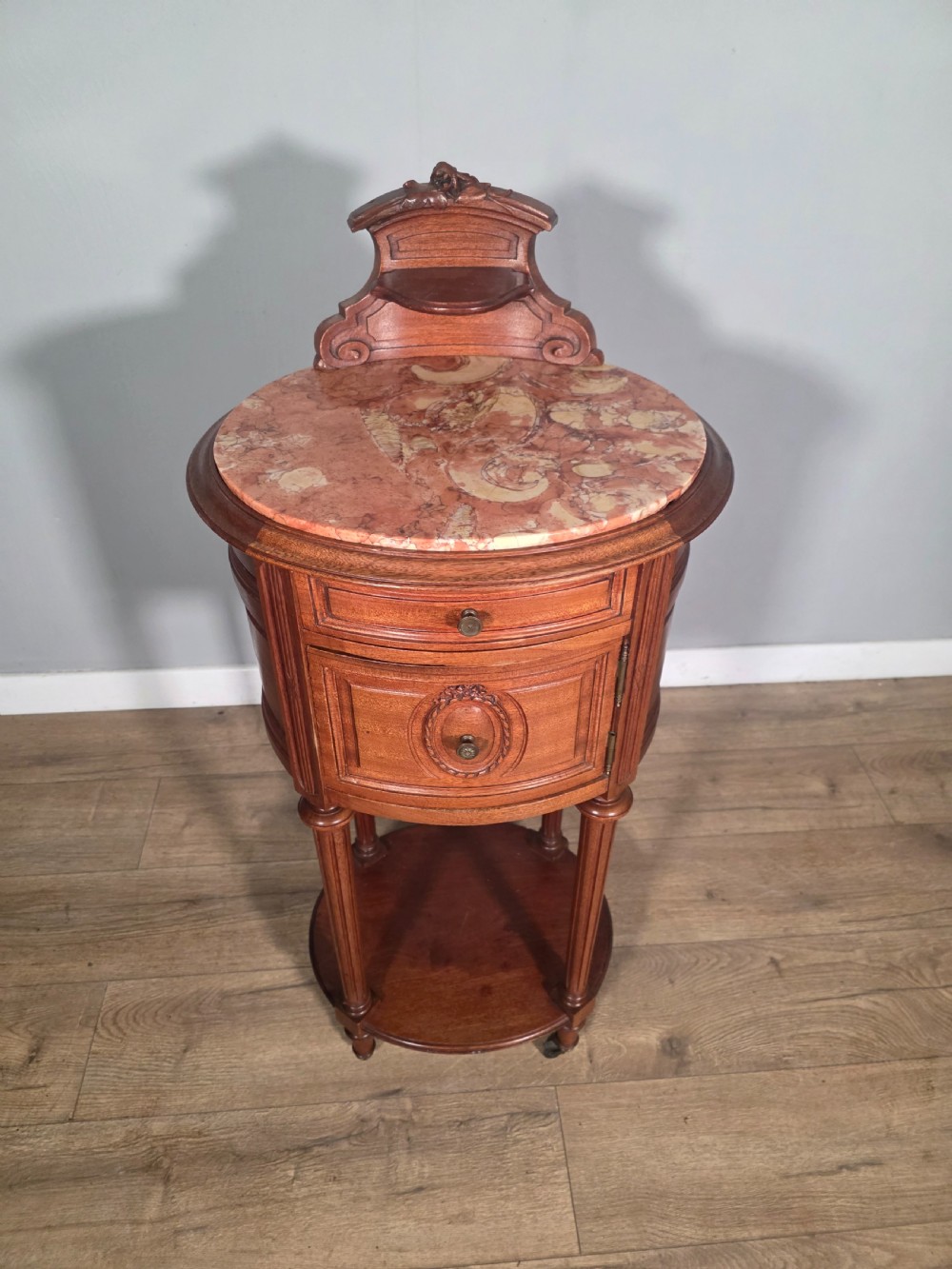 An antique French mahogany bedside cabinet featuring an oval marble top, a carved cresting gallery, a single drawer above a cupboard door, turned fluted legs, and a lower shelf. View IV