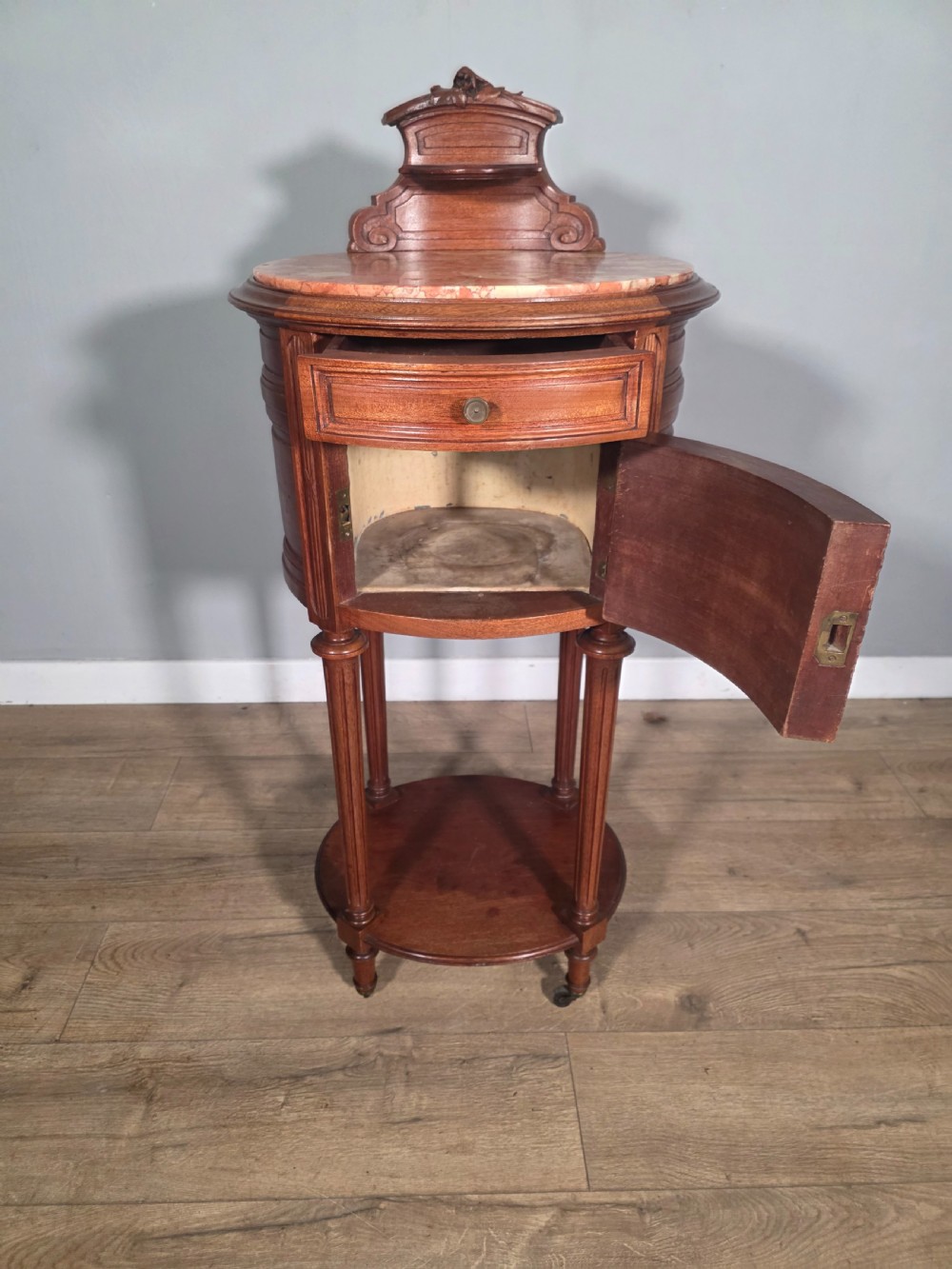 An antique French mahogany bedside cabinet featuring an oval marble top, a carved cresting gallery, a single drawer above a cupboard door, turned fluted legs, and a lower shelf. View V