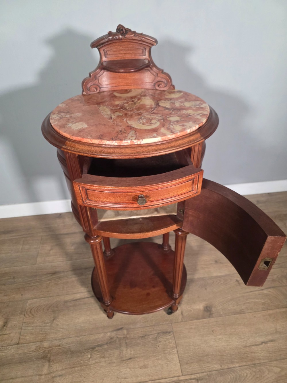 An antique French mahogany bedside cabinet featuring an oval marble top, a carved cresting gallery, a single drawer above a cupboard door, turned fluted legs, and a lower shelf. View VI