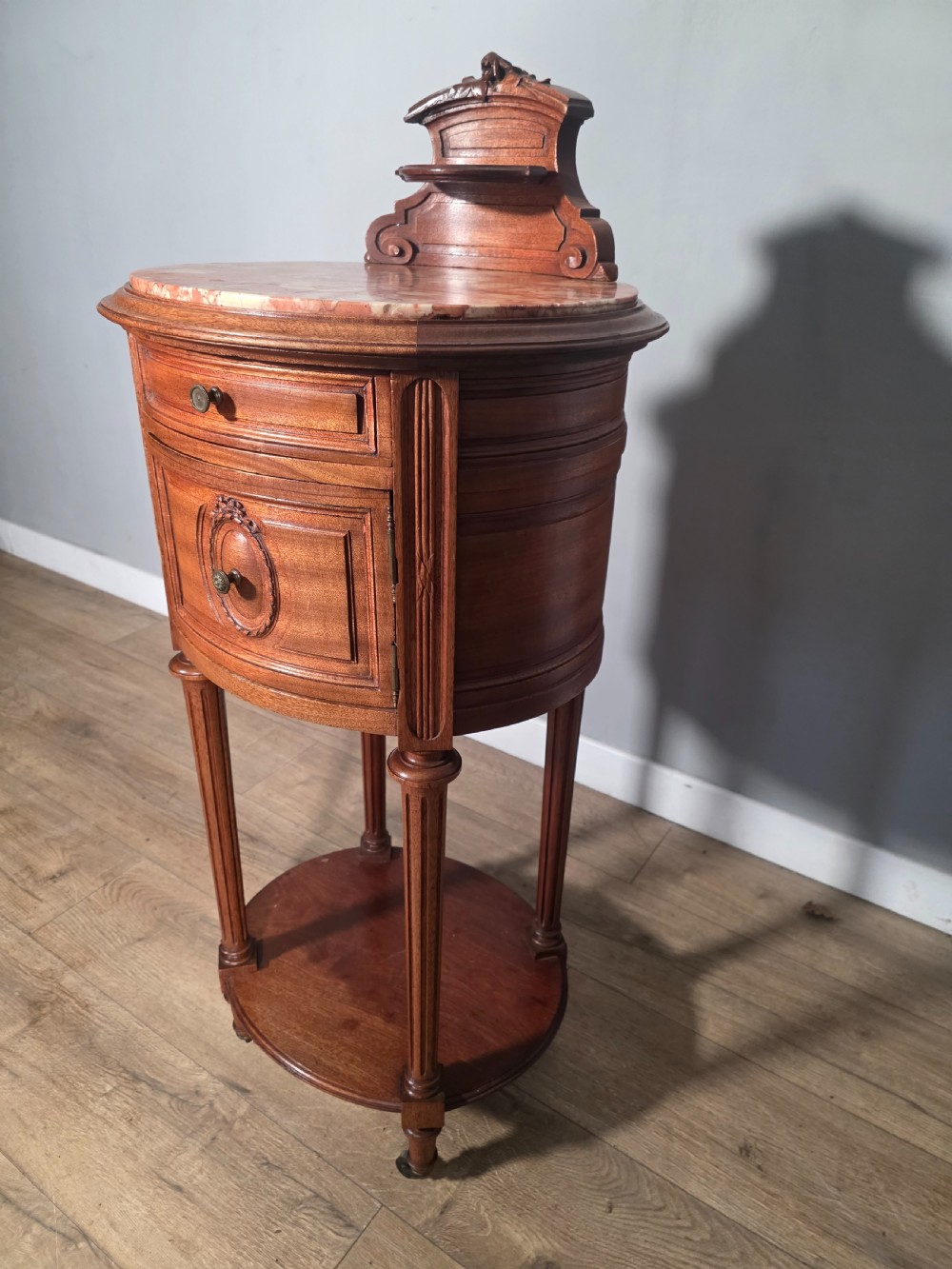 An antique French mahogany bedside cabinet featuring an oval marble top, a carved cresting gallery, a single drawer above a cupboard door, turned fluted legs, and a lower shelf. View VII