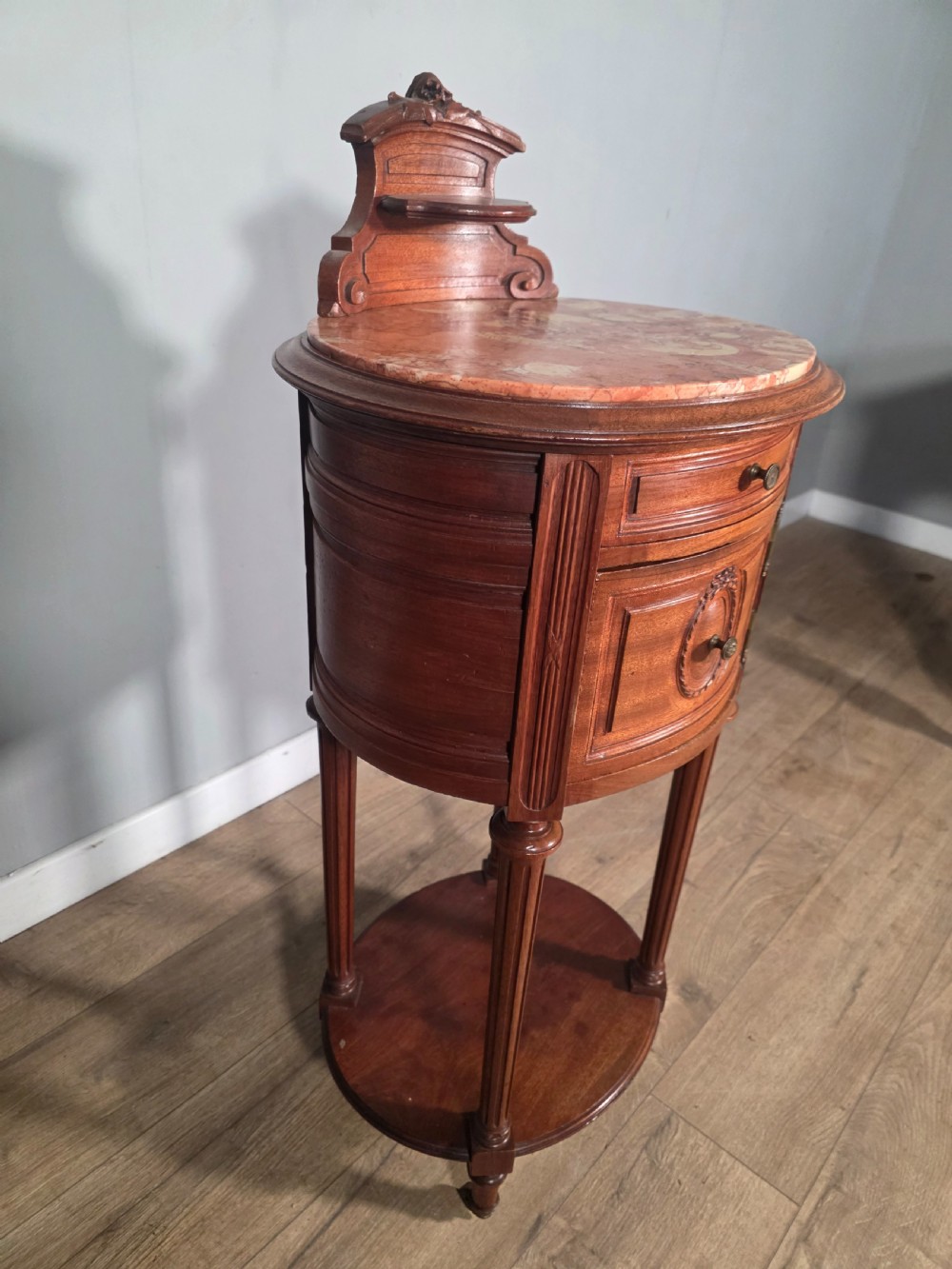 An antique French mahogany bedside cabinet featuring an oval marble top, a carved cresting gallery, a single drawer above a cupboard door, turned fluted legs, and a lower shelf. View VIII
